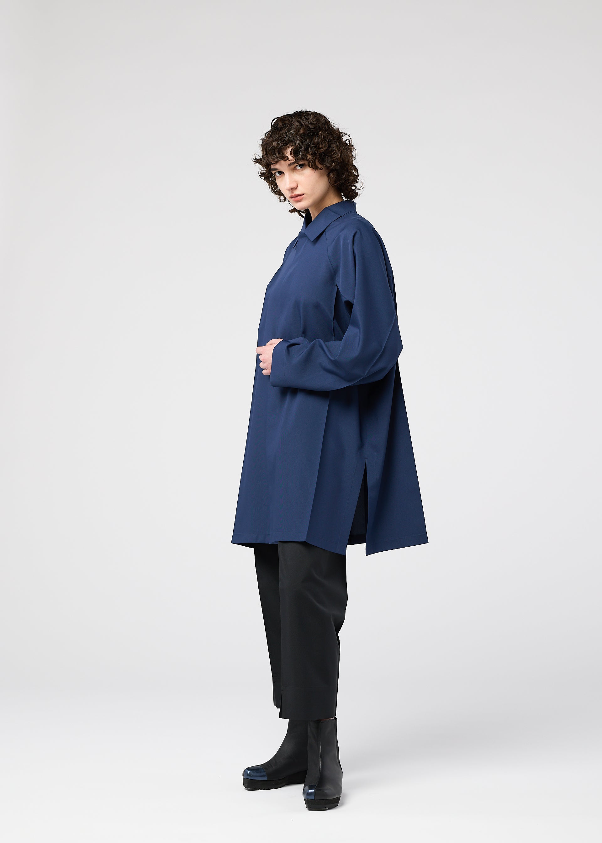 Zoomed view A person with curly hair stands in profile, wearing the oversized navy FOLD SHIRT by 132 5. ISSEY MIYAKE over black pants and black heeled ankle boots, set against a plain light-gray background.