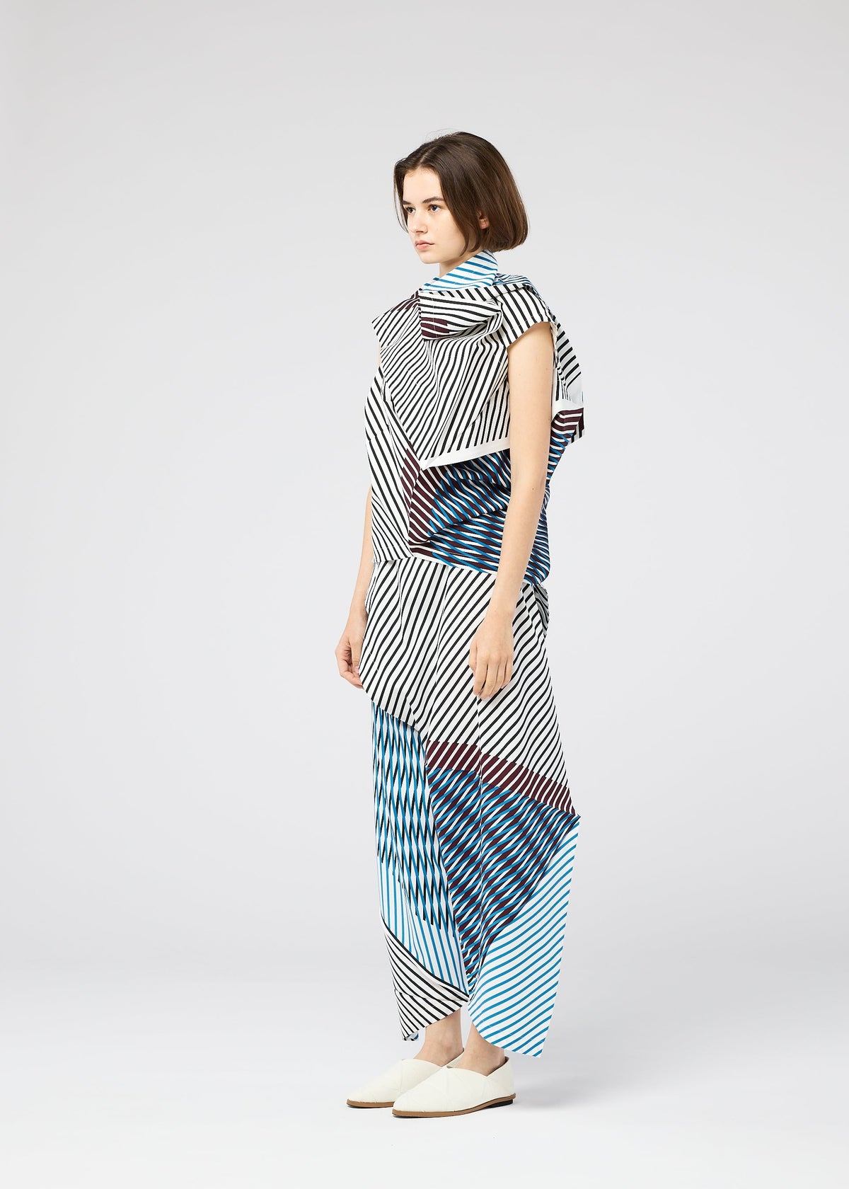 A woman in profile wears the FACET outfit by 132 5. ISSEY MIYAKE, featuring geometric black-and-white stripes and blue patterns. The Nassen print creates a striking, three-dimensional effect against a plain background. view 6 of 9
