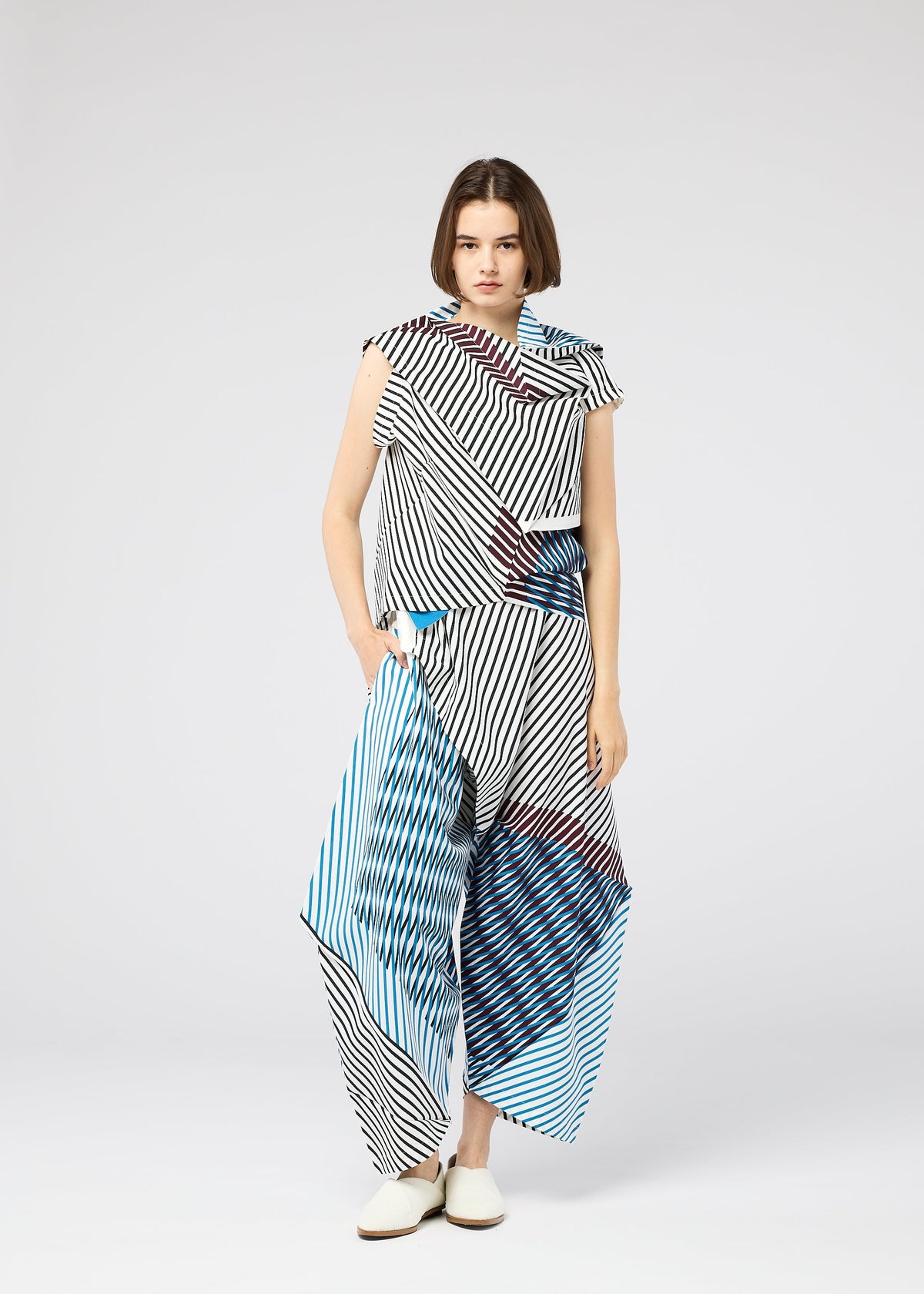 A woman with a bob stands against a plain background wearing the 132 5. ISSEY MIYAKE FACET black-and-white striped set with blue and red accents and white shoes, creating a bold, three-dimensional visual effect. view 9 of 9