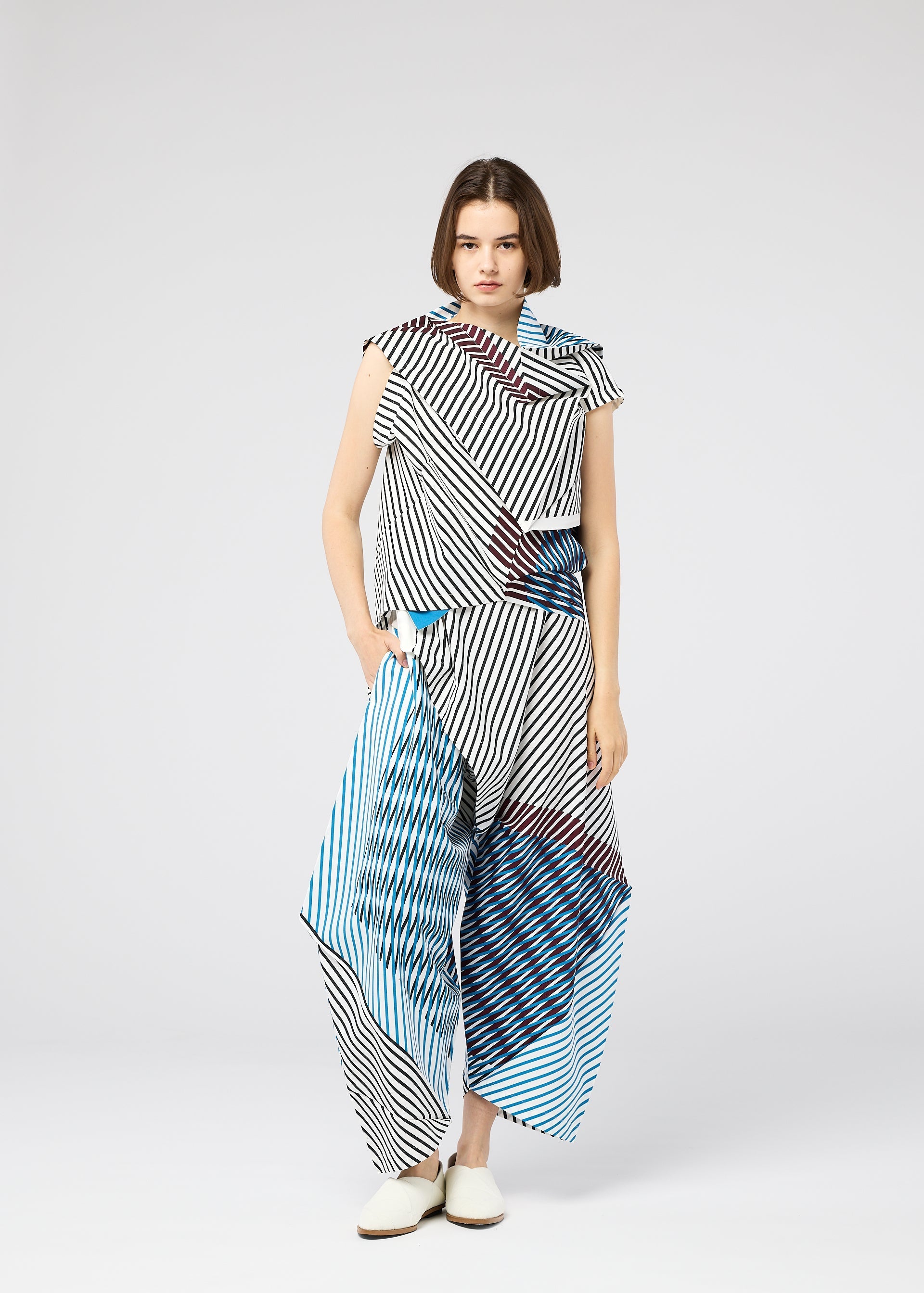 Zoomed view A woman with a bob stands against a plain background wearing the 132 5. ISSEY MIYAKE FACET black-and-white striped set with blue and red accents and white shoes, creating a bold, three-dimensional visual effect.