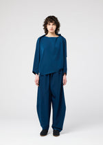 A person with curly hair faces forward, wearing the PARALLEL WRAP by 132 5. ISSEY MIYAKE—a dark blue, wrinkle-resistant top and wide-leg pants set in lightweight kersey—with black shoes against a plain light gray background.