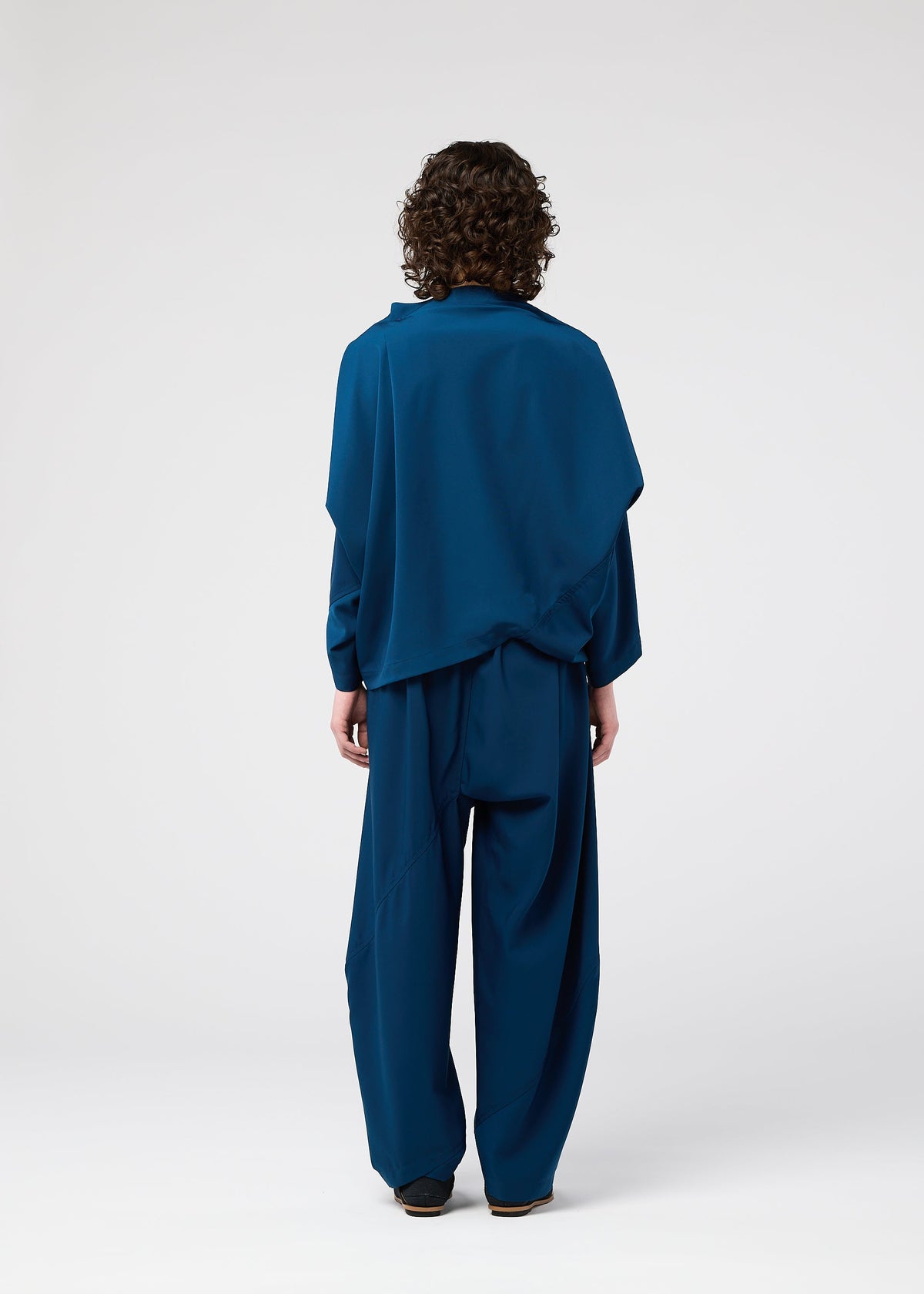 A person with curly hair stands facing away from the camera, wearing the PARALLEL WRAP by 132 5. ISSEY MIYAKE—a loose-fitting, blue, wrinkle-resistant top and wide-legged lightweight kersey pants—against a plain white background. view 7 of 9