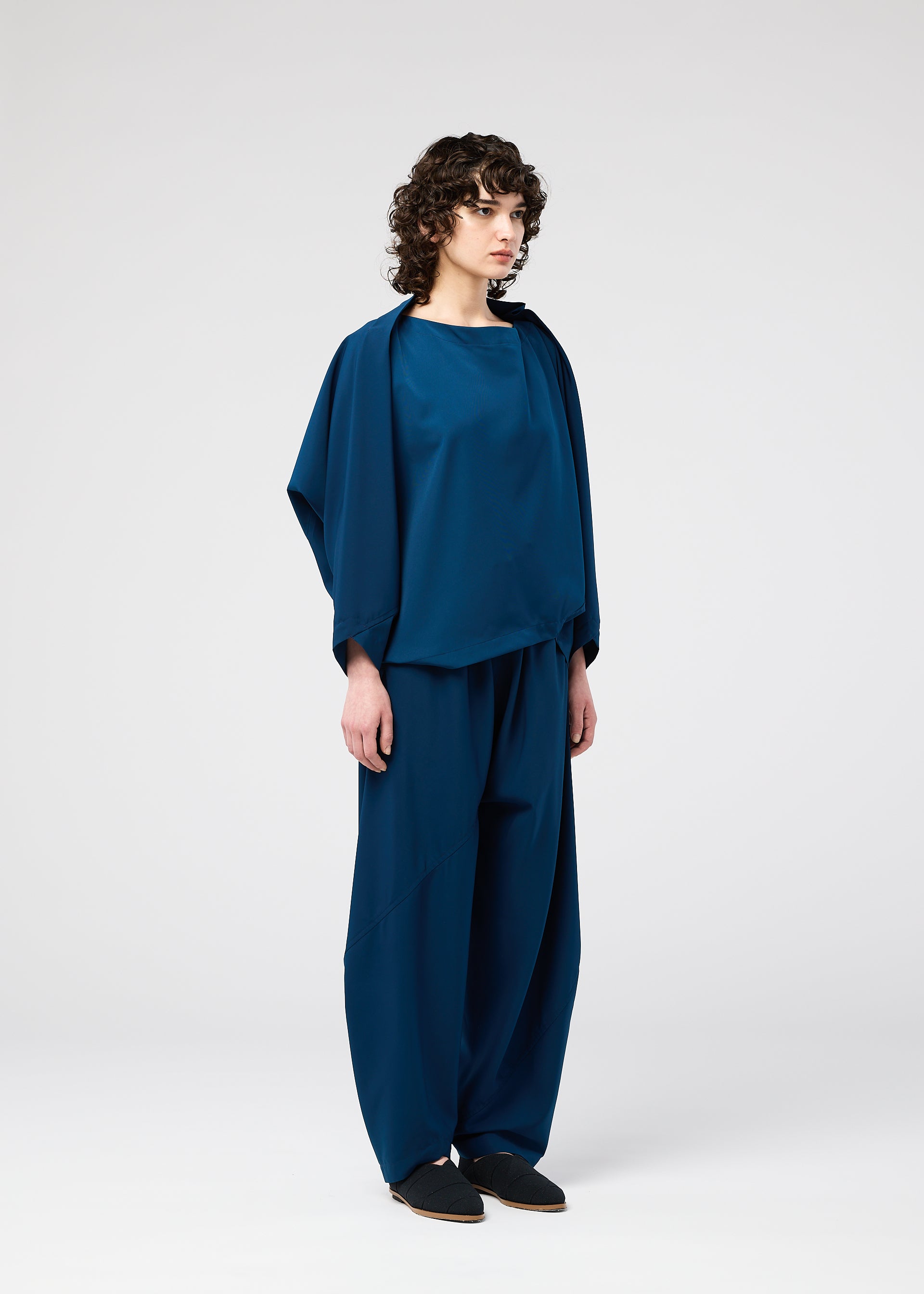 Zoomed view A person with curly hair stands in a studio wearing the loose-fitting PARALLEL WRAP by 132 5. ISSEY MIYAKE, paired with wide-leg pants and black slip-on shoes. The background is plain and light-colored.