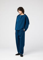 A person with curly hair stands against a white background, wearing the PARALLEL WRAP by 132 5. ISSEY MIYAKE—a loose-fitting, wrinkle-resistant top with matching blue kersey pants—paired with dark slip-on shoes.
