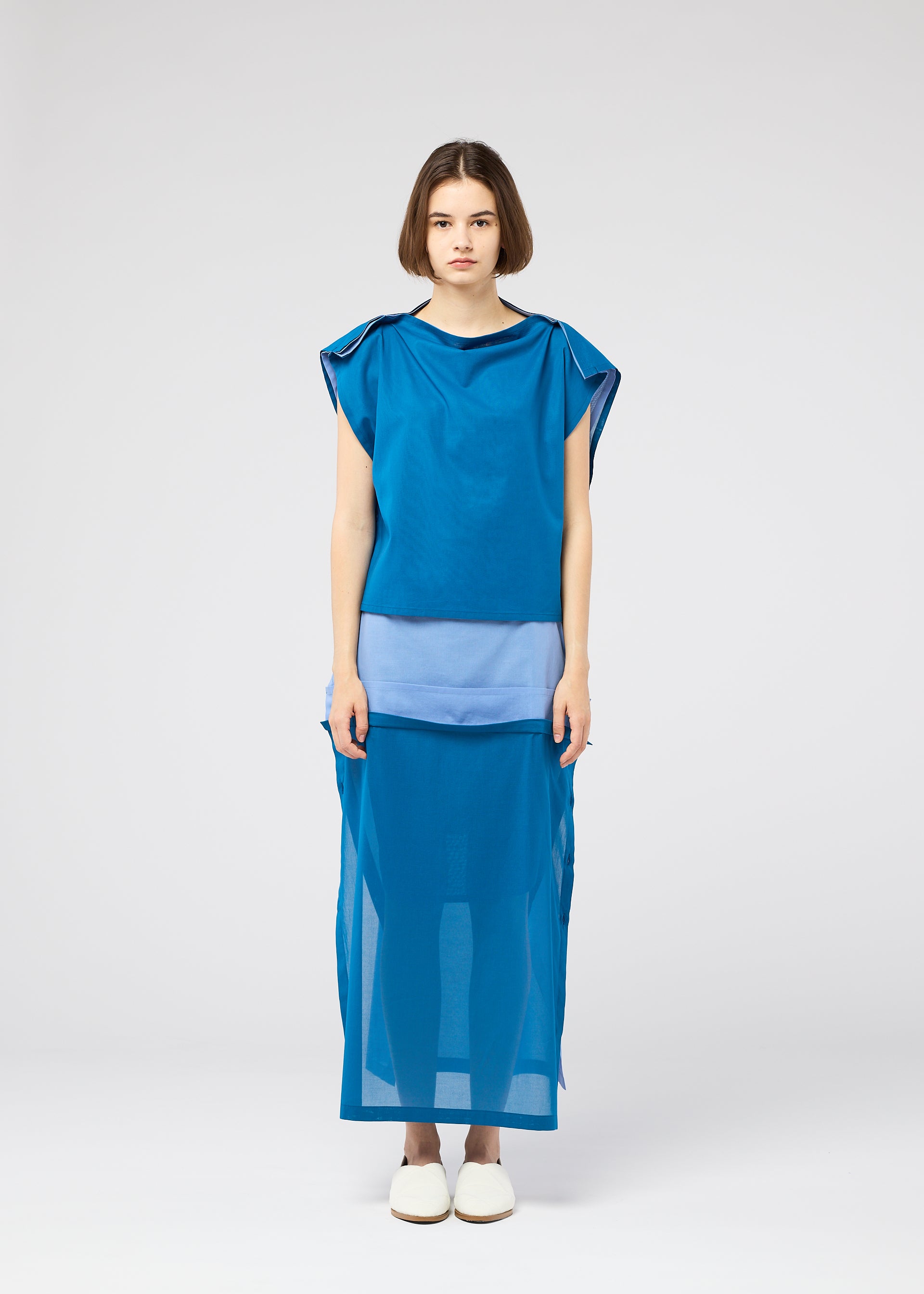 Zoomed view A woman with short brown hair faces forward wearing the SHEER FOLIO loose-fit blue dress by 132 5. ISSEY MIYAKE, featuring fabric layers and sheer panels over a light blue petticoat, paired with white slip-on shoes against a plain white background.