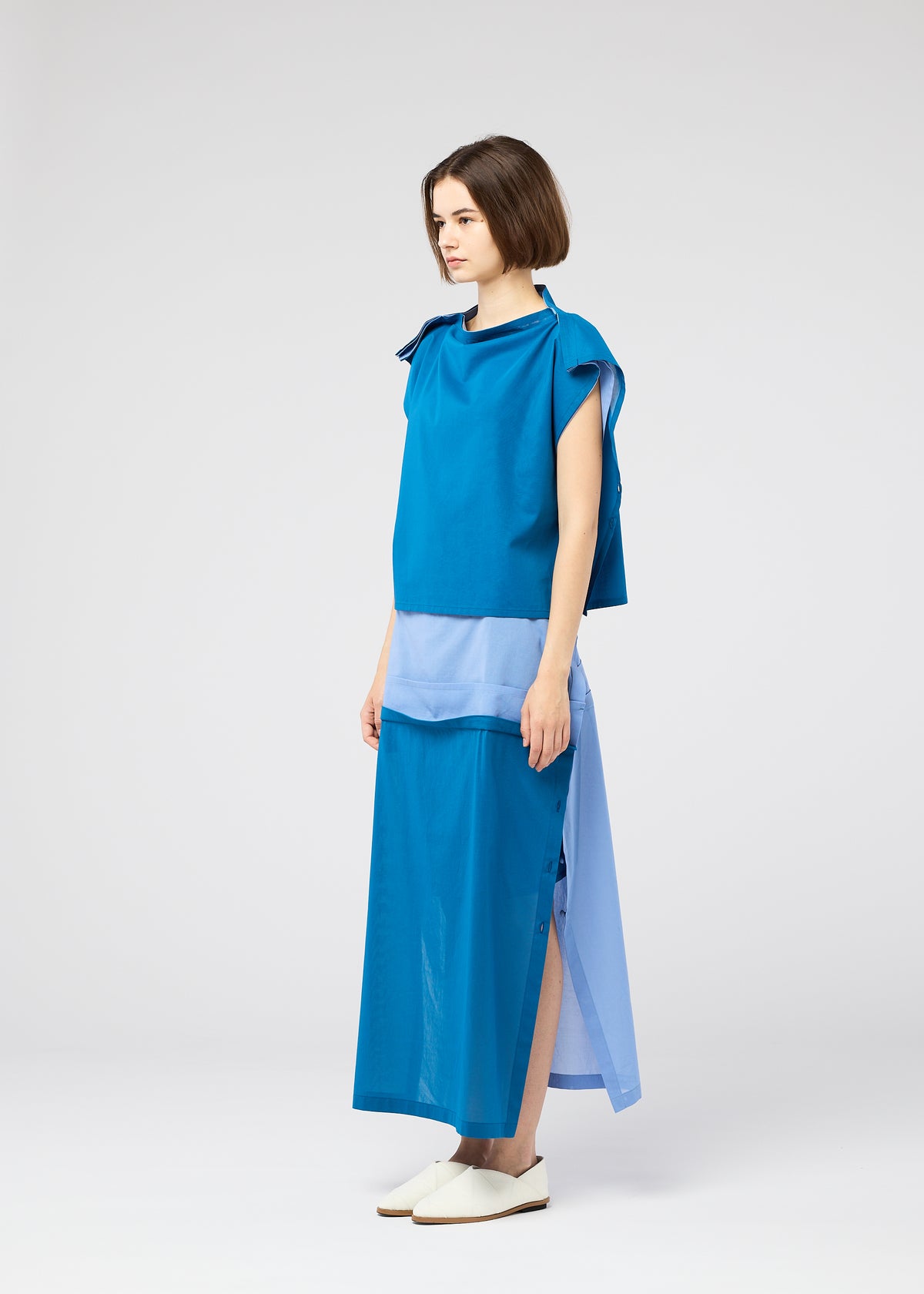 A woman with short brown hair wears the SHEER FOLIO by 132 5. ISSEY MIYAKE: a sleeveless blue top and long, layered blue skirt in various shades, paired with white slip-on shoes, standing sideways against a plain light background. view 7 of 10