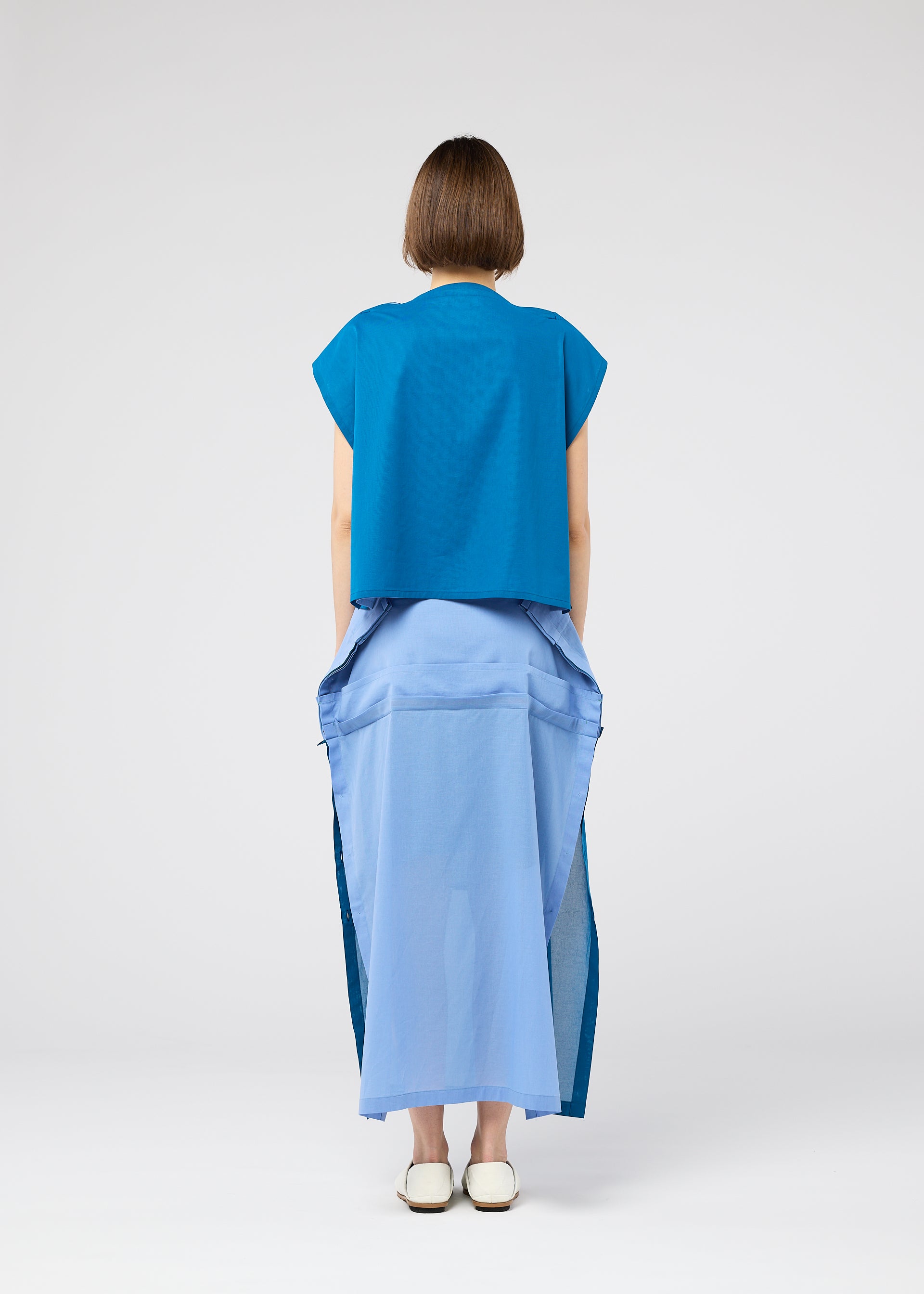 Zoomed view A person with short brown hair faces away, wearing the SHEER FOLIO dress by 132 5. ISSEY MIYAKE—blue top, layered light blue dress with side slits, and white shoes—set against a simple, light background.