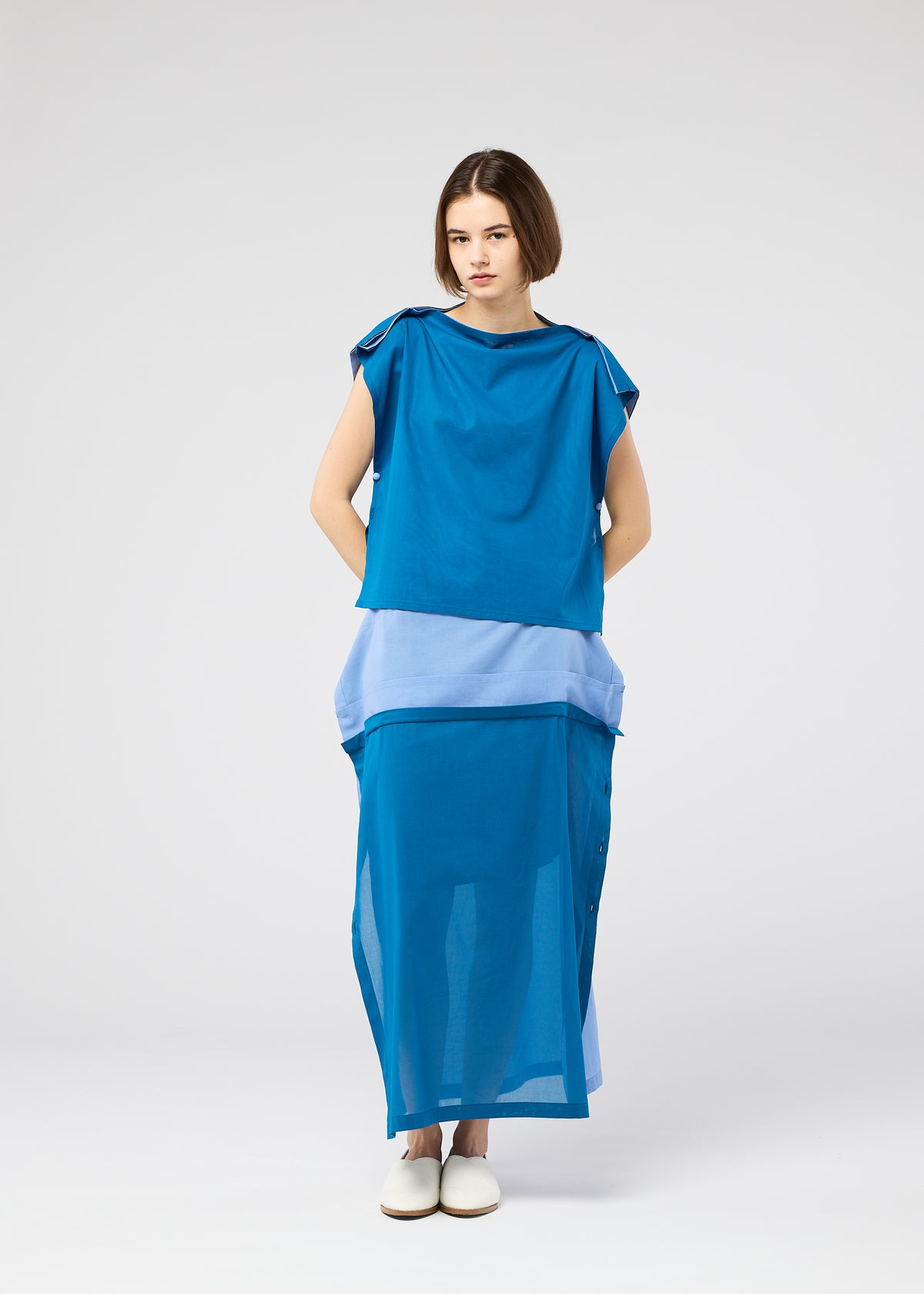 A woman with short brown hair and a neutral expression wears the SHEER FOLIO dress by 132 5. ISSEY MIYAKE—a loose, sleeveless blue piece with layered fabric and lighter blue bands at the waist and hem—paired with white slip-on shoes. view 9 of 10