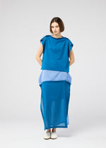 A woman with short brown hair and a neutral expression wears the SHEER FOLIO dress by 132 5. ISSEY MIYAKE—a loose, sleeveless blue piece with layered fabric and lighter blue bands at the waist and hem—paired with white slip-on shoes.