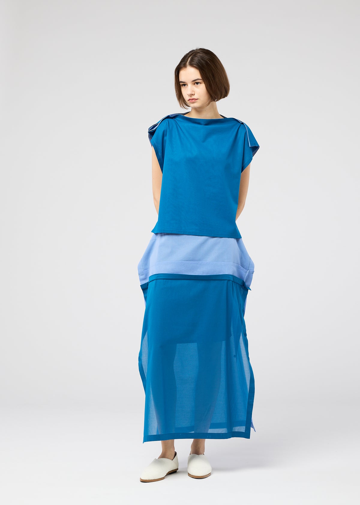 A woman with short brown hair and white slip-on shoes stands against a plain light background, wearing the SHEER FOLIO blue and light blue layered dress by 132 5. ISSEY MIYAKE, featuring a sheer bottom section. view 10 of 10