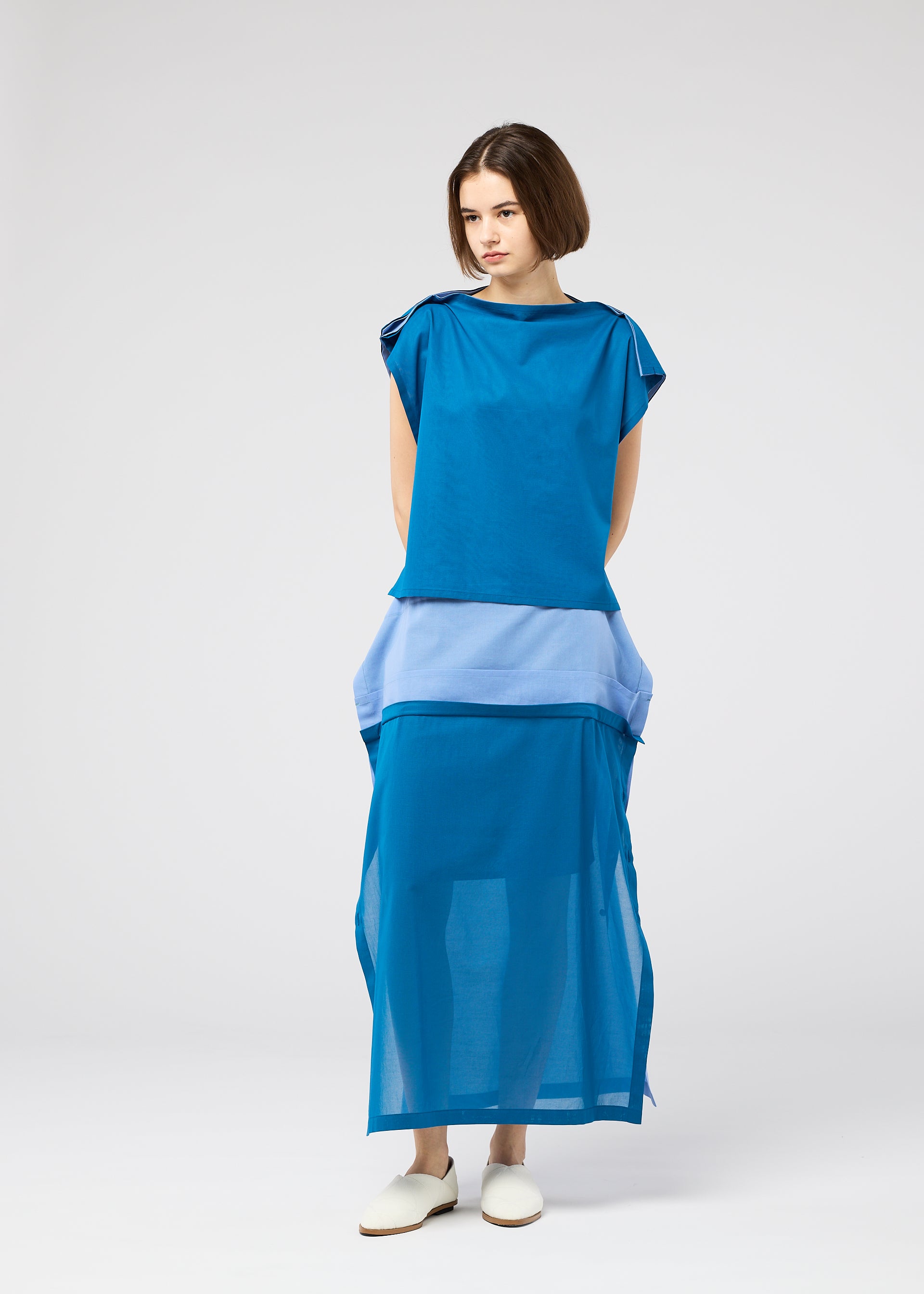 Zoomed view A woman with short brown hair and white slip-on shoes stands against a plain light background, wearing the SHEER FOLIO blue and light blue layered dress by 132 5. ISSEY MIYAKE, featuring a sheer bottom section.