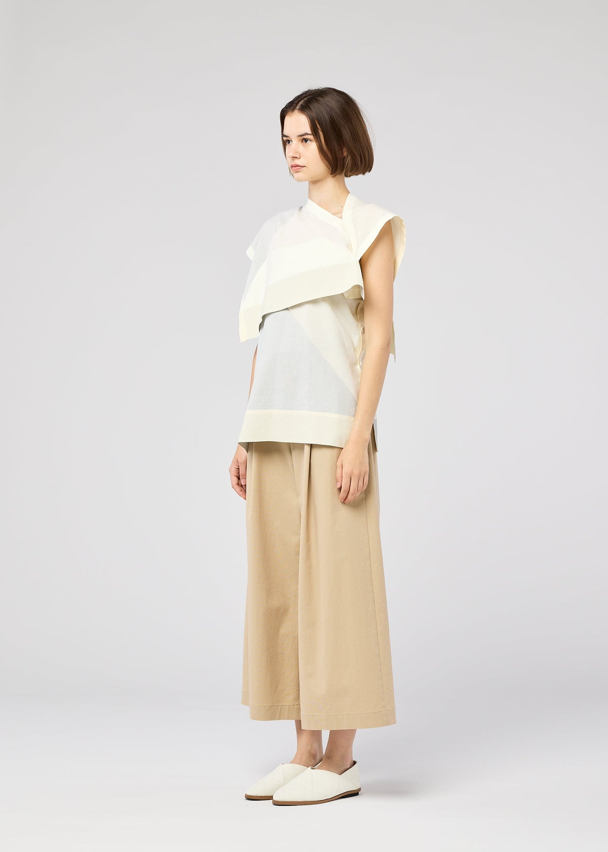 A woman with short brown hair faces left, wearing the FOLD RAMIE PRINT white sleeveless top by 132 5. ISSEY MIYAKE, paired with wide beige cropped pants and white slip-on shoes, set against a plain light background. view 8 of 11