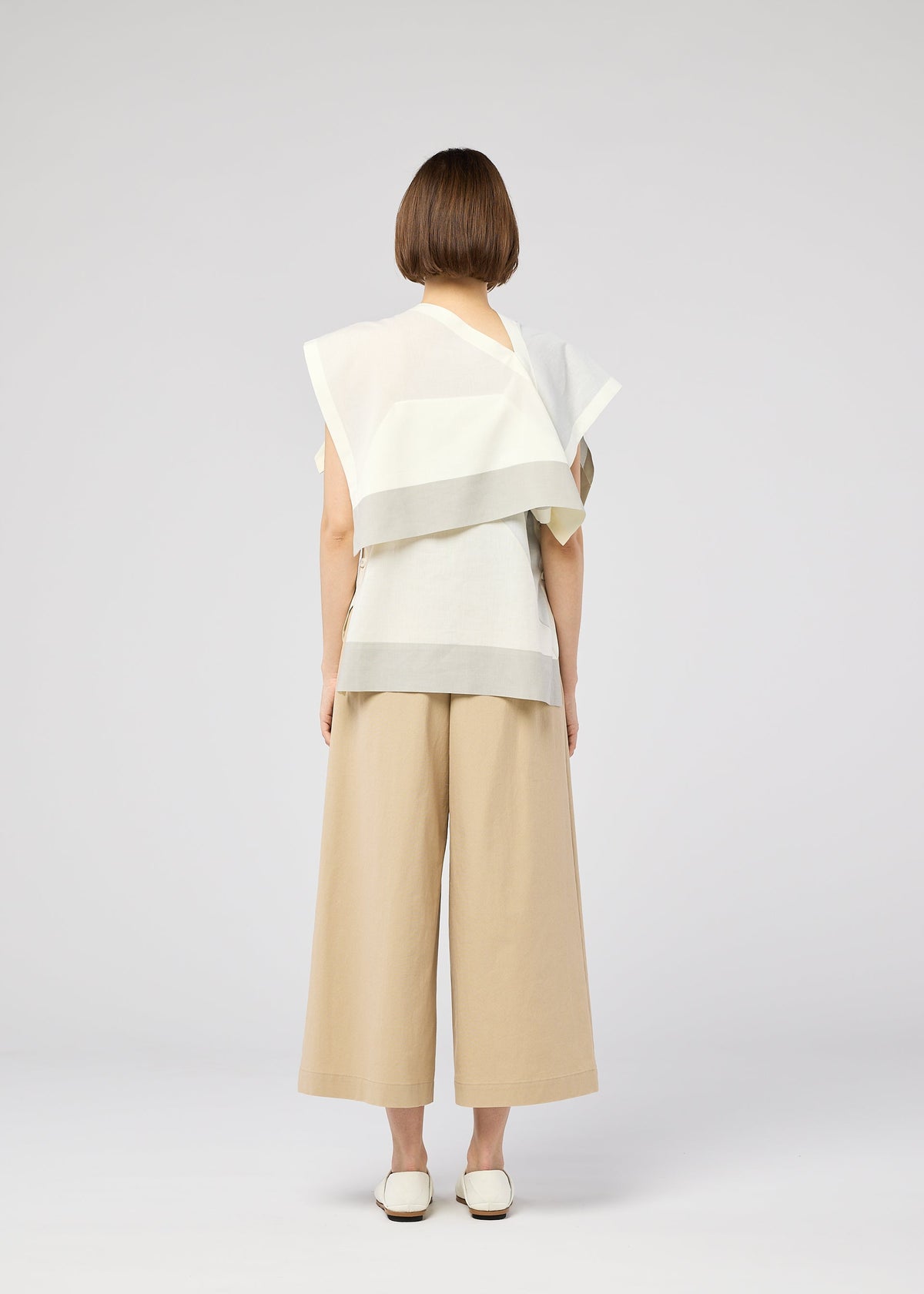 A person with short brown hair faces away, wearing the FOLD RAMIE PRINT top by 132 5. ISSEY MIYAKE, paired with wide-leg beige pants and white shoes against a plain light gray background. view 9 of 11
