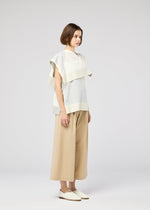 A person with a bob stands sideways, wearing the FOLD RAMIE PRINT top by 132 5. ISSEY MIYAKE—loose, layered white and light gray with a subtle print—paired with wide beige pants and white slip-ons against a plain light gray background.