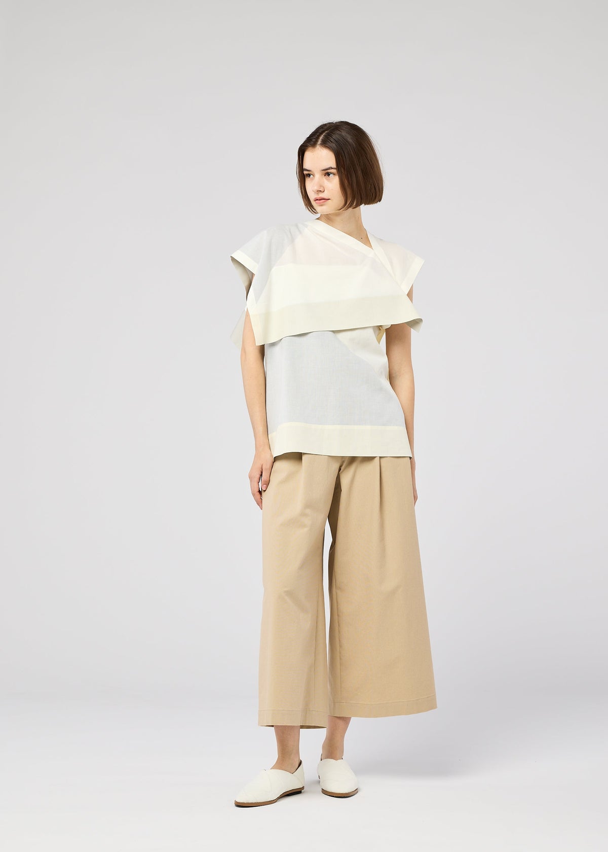 A woman with a bob stands against a plain light gray background, wearing the 132 5. ISSEY MIYAKE FOLD RAMIE PRINT top in cream and light blue, wide beige culottes, and white slip-on shoes. view 11 of 11