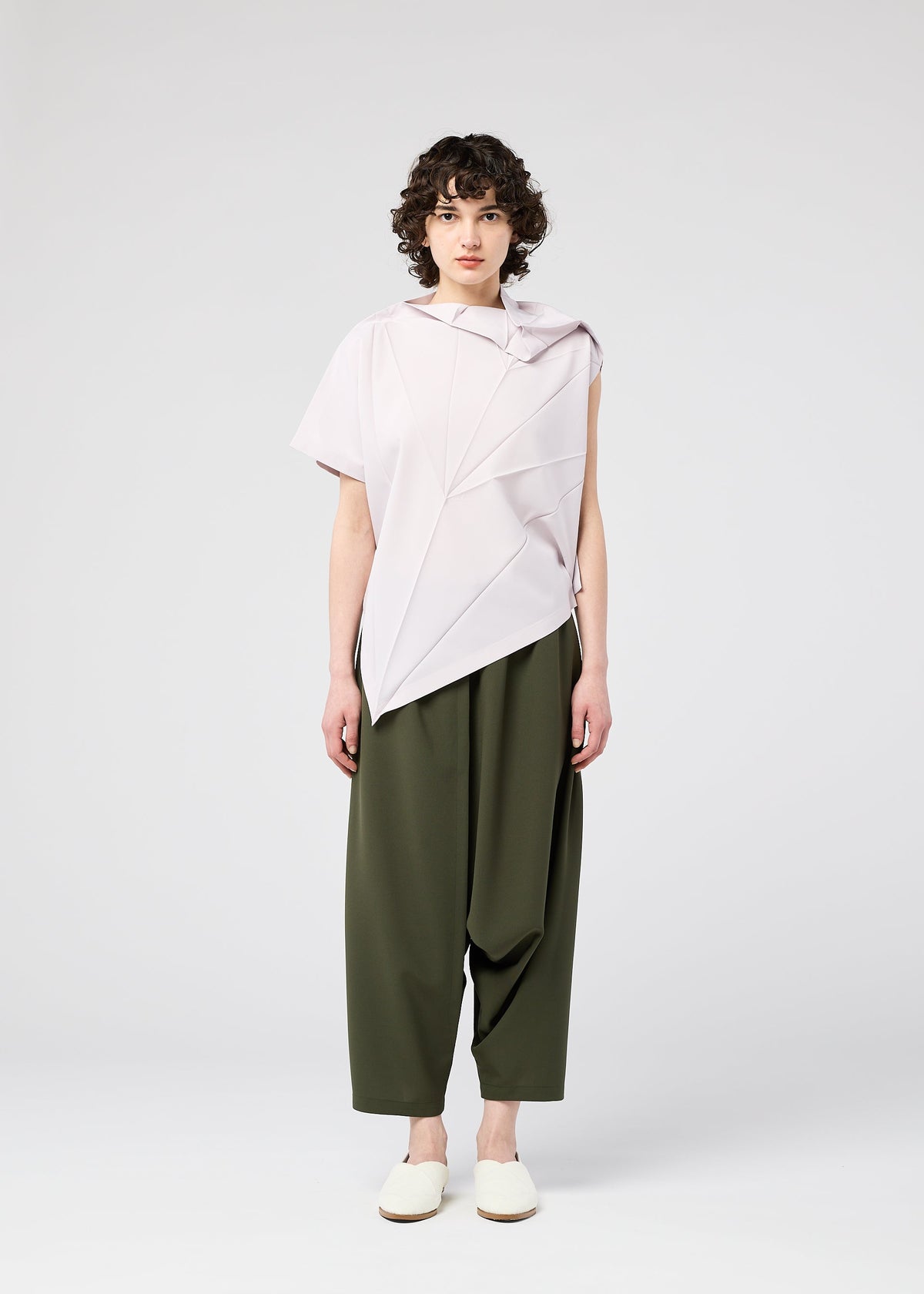 Against a light gray background, a person with short, curly hair wears the 132 5. ISSEY MIYAKE RADIAL FOLD SHIRT in light pink, loose olive green pants, and white slip-on shoes. view 8 of 12