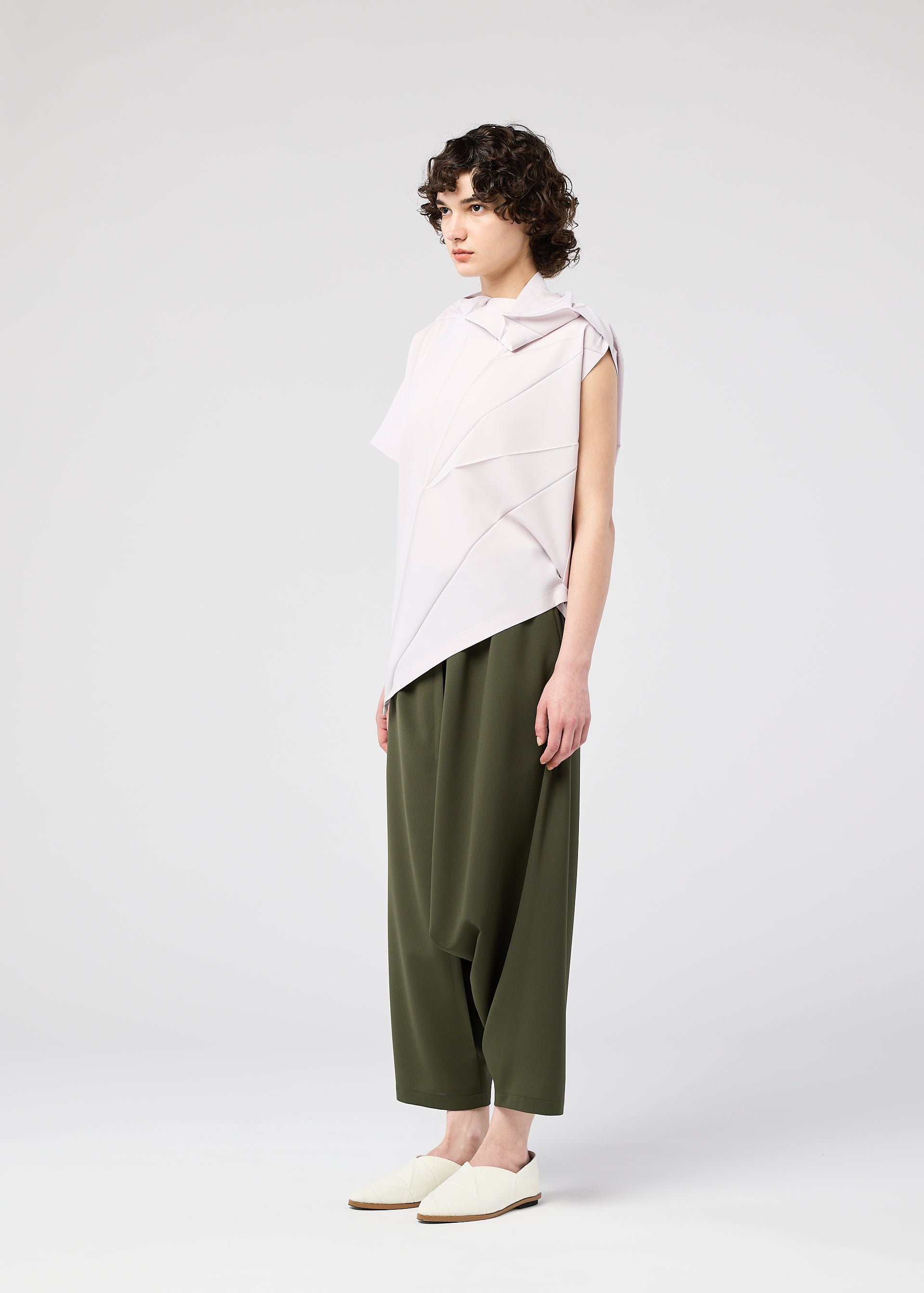 Zoomed view A person with short curly hair models the 132 5. ISSEY MIYAKE RADIAL FOLD SHIRT, paired with loose olive green pants and white slip-on shoes, standing against a plain background with a neutral expression.