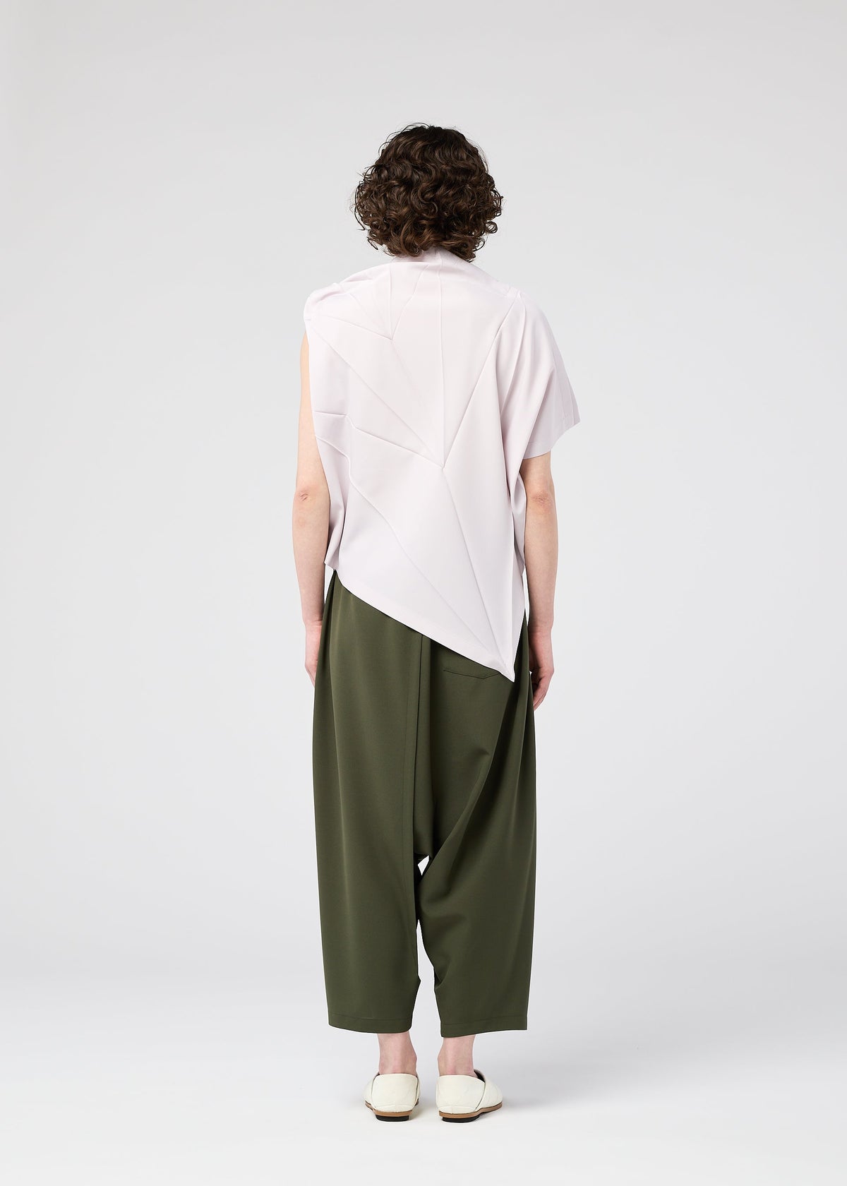Seen from behind, a person with short curly hair wears the 132 5. ISSEY MIYAKE RADIAL FOLD SHIRT, loose olive green pants, and white flat shoes against a plain light gray background. view 10 of 12