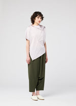 A person with short curly hair wears the RADIAL FOLD SHIRT by 132 5. ISSEY MIYAKE, featuring a light, draped asymmetrical design and comfortable fabric, styled with loose olive green pants and slip-on shoes against a white background.