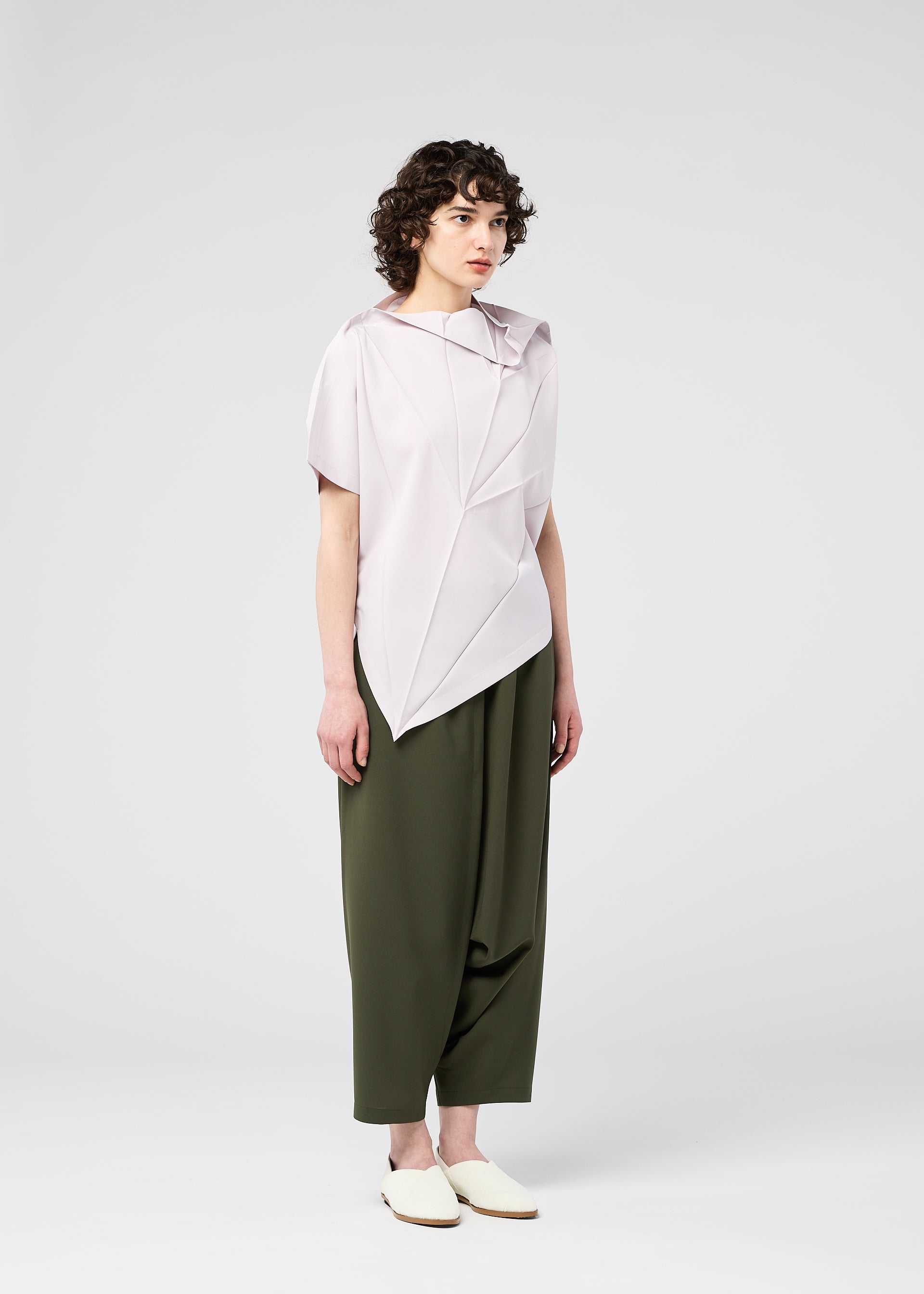 Zoomed view A person with short curly hair wears the RADIAL FOLD SHIRT by 132 5. ISSEY MIYAKE, featuring a light, draped asymmetrical design and comfortable fabric, styled with loose olive green pants and slip-on shoes against a white background.