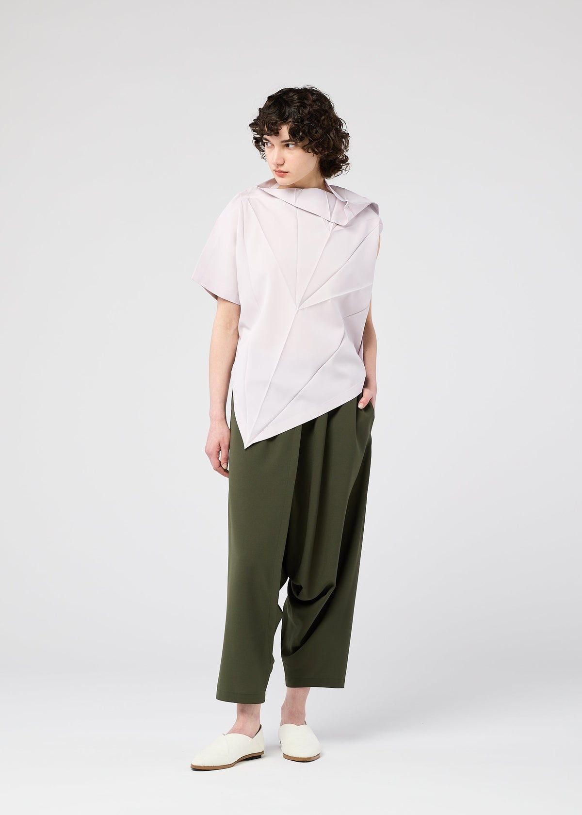 A person with short, curly hair wears the 132 5. ISSEY MIYAKE RADIAL FOLD SHIRT in light pink, featuring a structured collar and comfortable fabric, paired with loose olive green pants against a plain white background. view 12 of 12