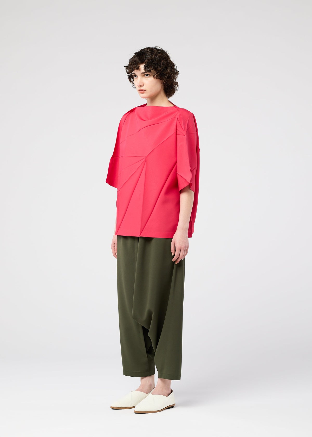 A person with short curly hair stands against a plain background, wearing the bright pink RADIAL FOLD SHIRT by 132 5. ISSEY MIYAKE with wide sleeves, paired with olive green loose pants and white slip-on shoes. view 8 of 11