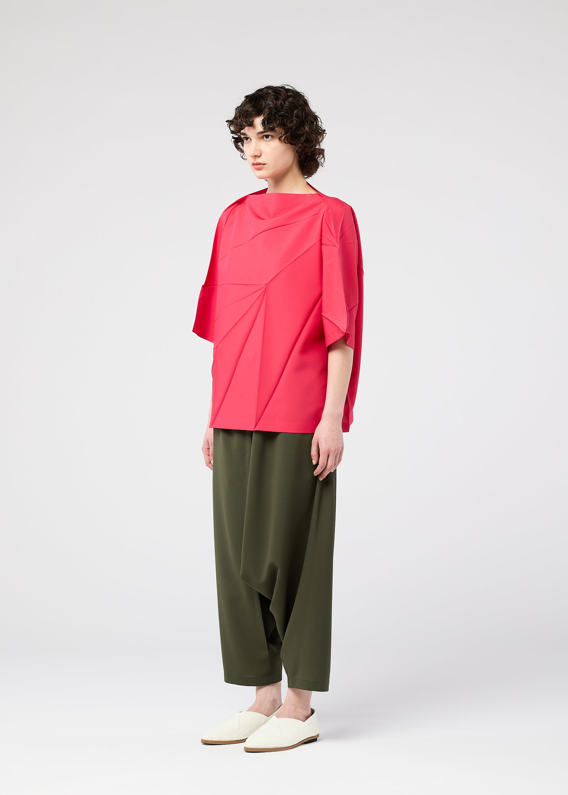Zoomed view A person with short curly hair stands against a plain background, wearing the bright pink RADIAL FOLD SHIRT by 132 5. ISSEY MIYAKE with wide sleeves, paired with olive green loose pants and white slip-on shoes.