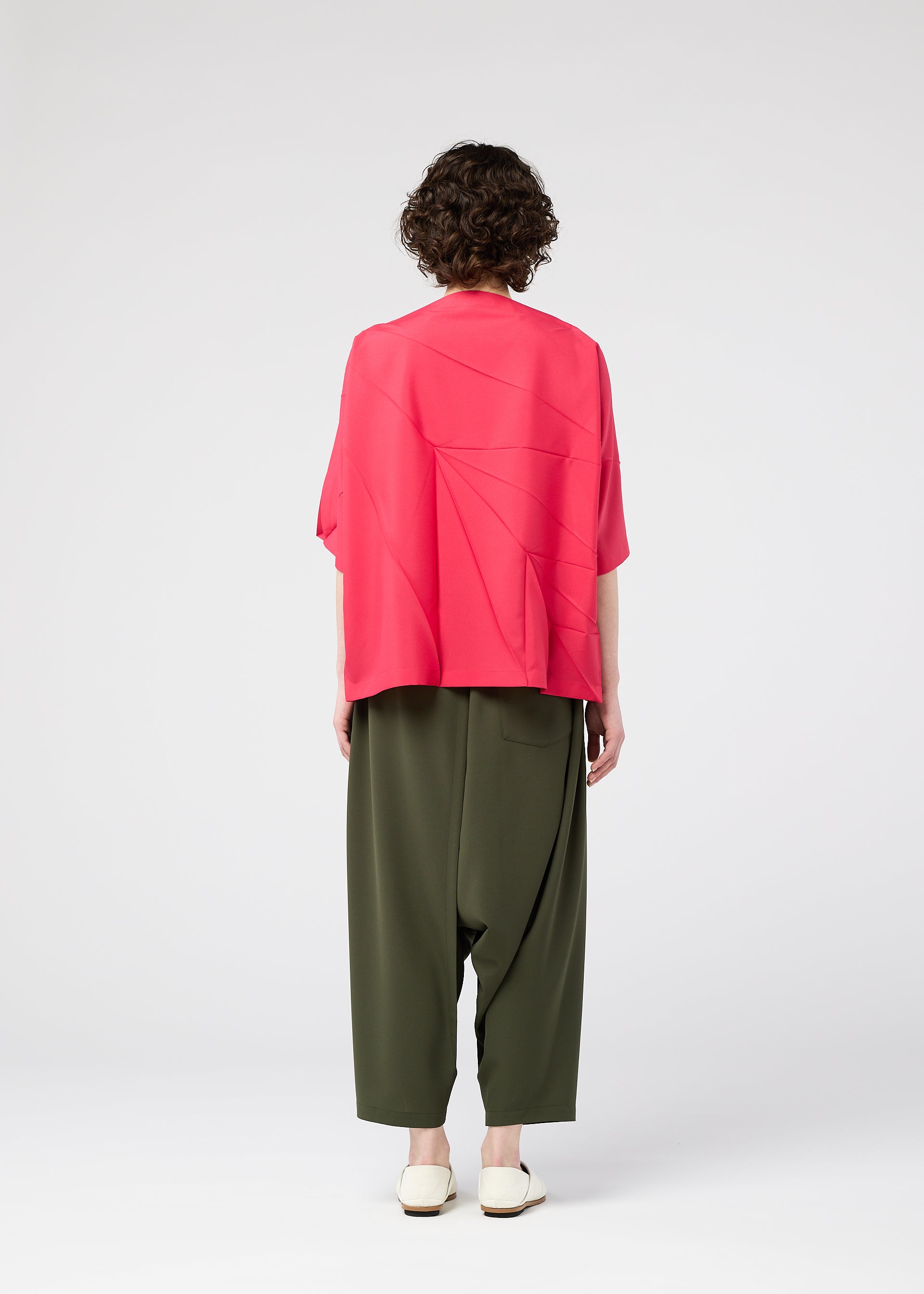 Zoomed view A person with short, curly brown hair stands facing away, wearing the 132 5. ISSEY MIYAKE RADIAL FOLD SHIRT in bright pink, baggy olive green pants, and white shoes against a plain white background.
