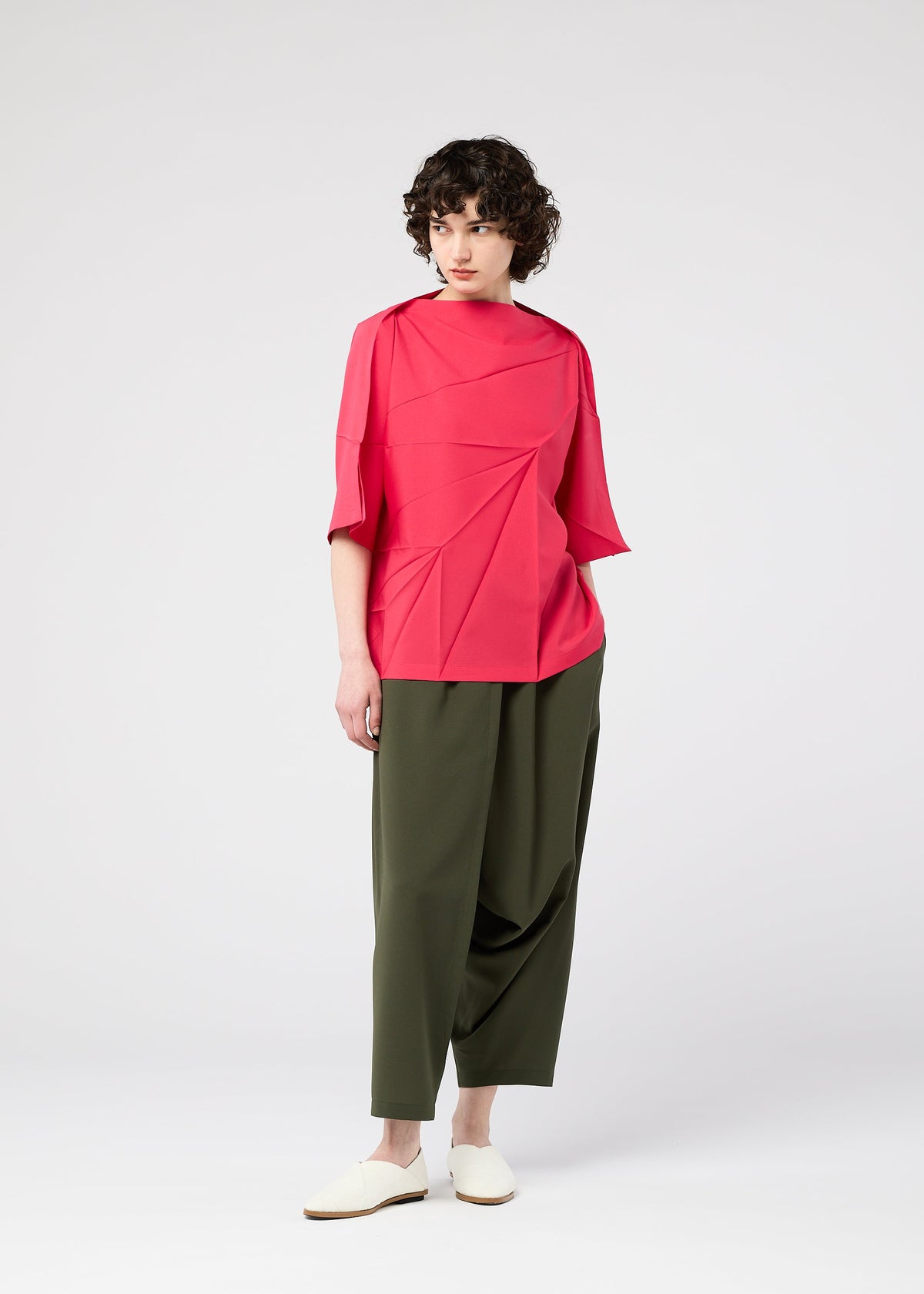 A person with short curly hair wears the RADIAL FOLD SHIRT from 132 5. ISSEY MIYAKE, a bright pink geometric top, paired with olive green wide-leg pants and white slip-on shoes, standing against a plain white background. view 10 of 11