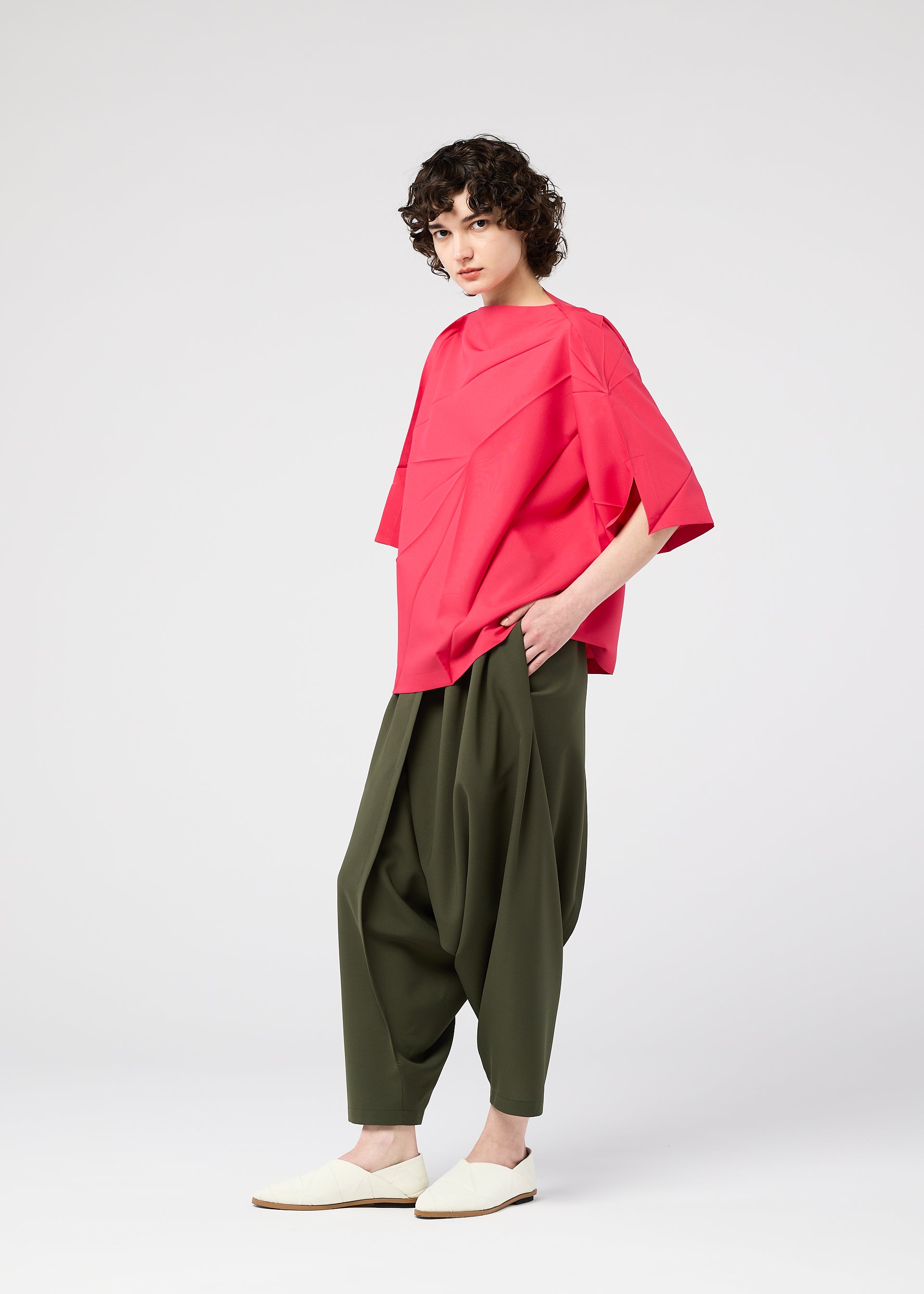 Zoomed view A person with short curly hair wears the loose, bright pink RADIAL FOLD SHIRT by 132 5. ISSEY MIYAKE, baggy olive-green pants, and cream slip-on shoes, standing against a plain white background with one hand in their pocket.