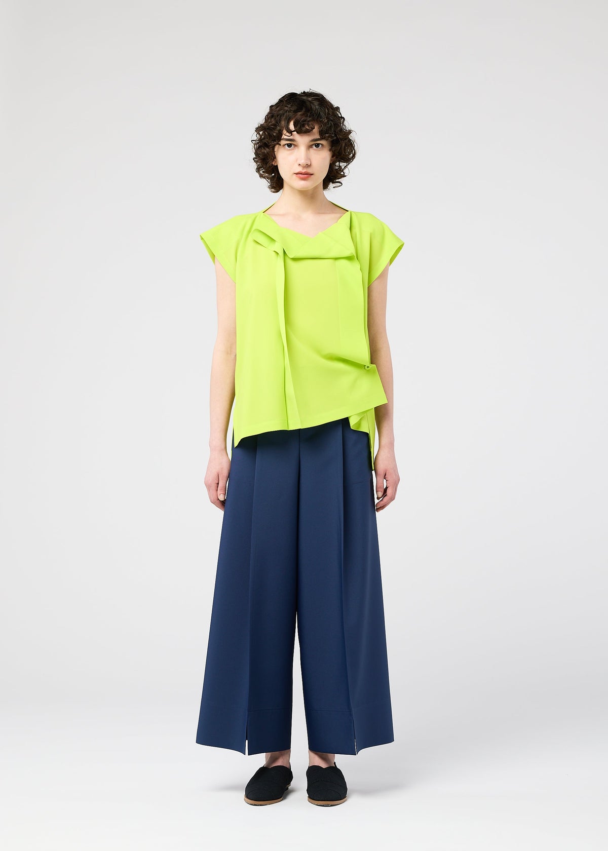 Against a plain light background, a person with short curly hair and a neutral expression wears the 132 5. ISSEY MIYAKE FLAT TUCK T in bright lime green, front-tucked, paired with wide-leg navy pants and black slip-on shoes. view 9 of 12