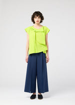 Against a plain light background, a person with short curly hair and a neutral expression wears the 132 5. ISSEY MIYAKE FLAT TUCK T in bright lime green, front-tucked, paired with wide-leg navy pants and black slip-on shoes.