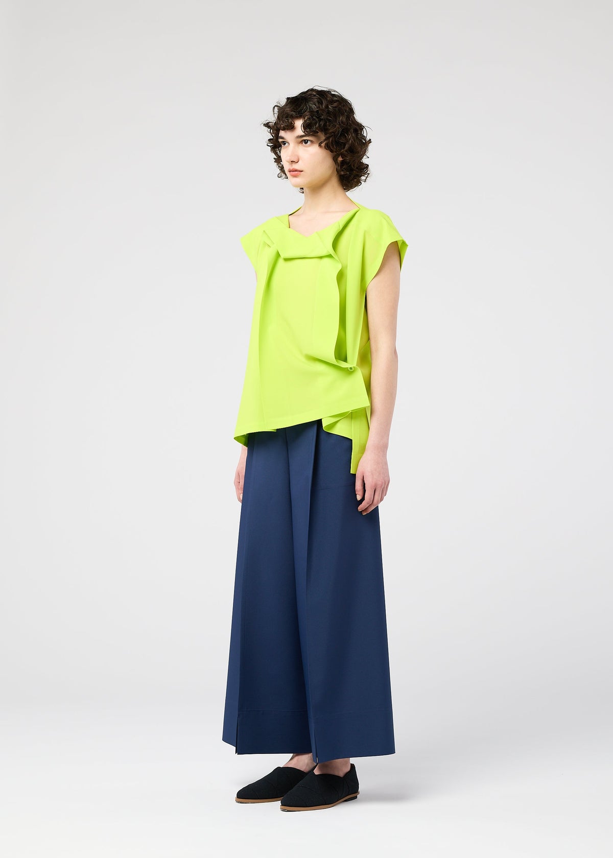 A person with short curly hair wears the FLAT TUCK T by 132 5. ISSEY MIYAKE in bright lime green, styled with wide-leg navy trousers and black shoes, standing against a plain white background. view 10 of 12