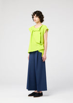 A person with short curly hair wears the FLAT TUCK T by 132 5. ISSEY MIYAKE in bright lime green, styled with wide-leg navy trousers and black shoes, standing against a plain white background.