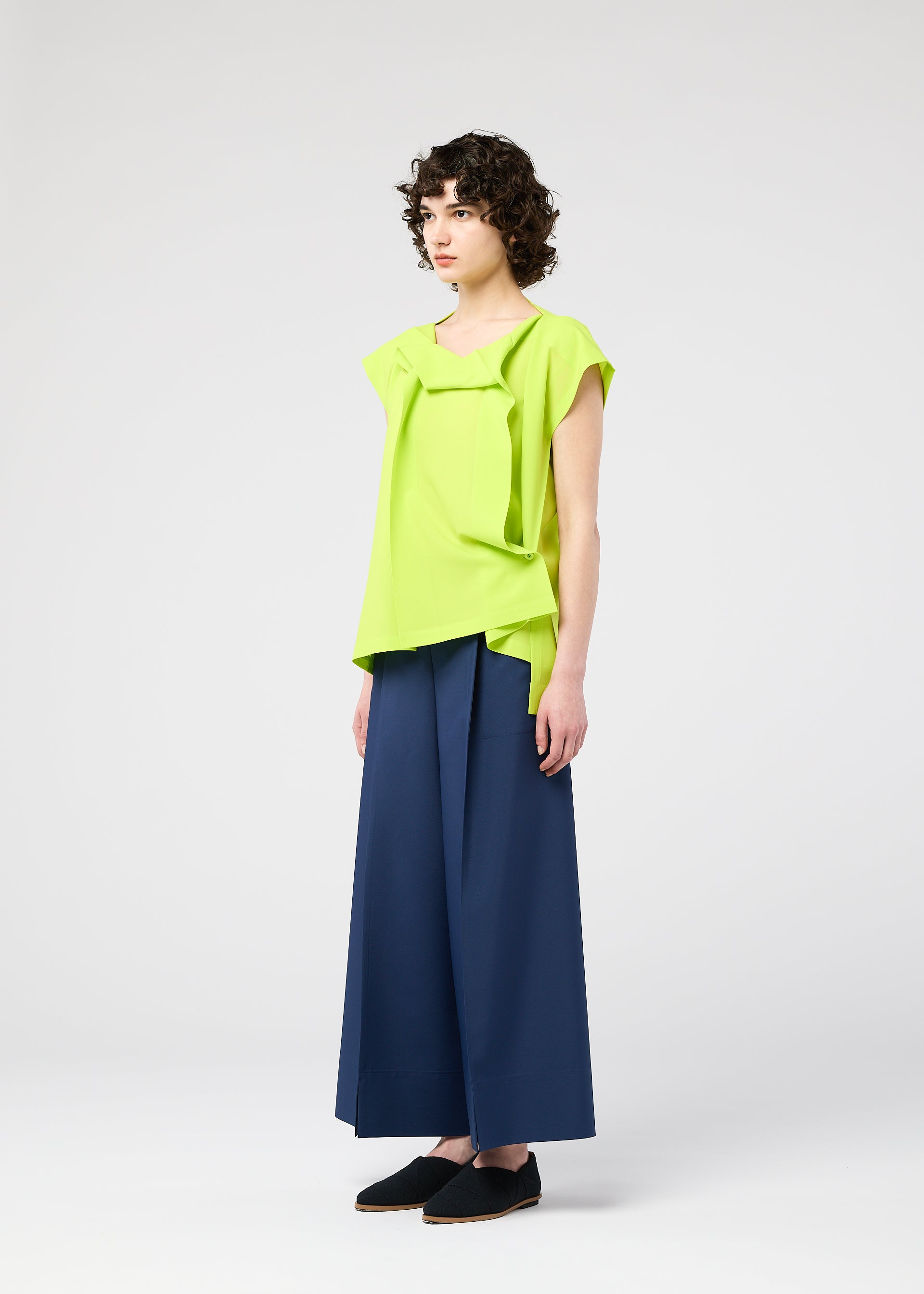 Zoomed view A person with short curly hair wears the FLAT TUCK T by 132 5. ISSEY MIYAKE in bright lime green, styled with wide-leg navy trousers and black shoes, standing against a plain white background.