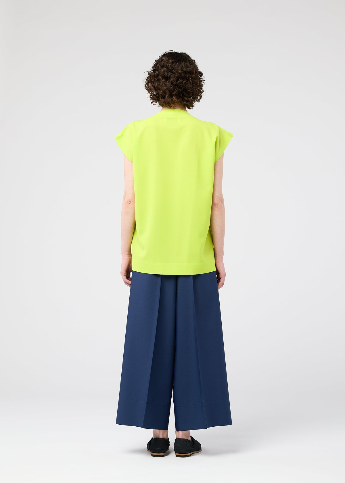 A person with short curly hair, seen from behind, wears the FLAT TUCK T by 132 5. ISSEY MIYAKE in bright yellow-green, styled front-tucked with wide-legged dark blue pants and black shoes against a light background for a gorgeous look. view 11 of 12