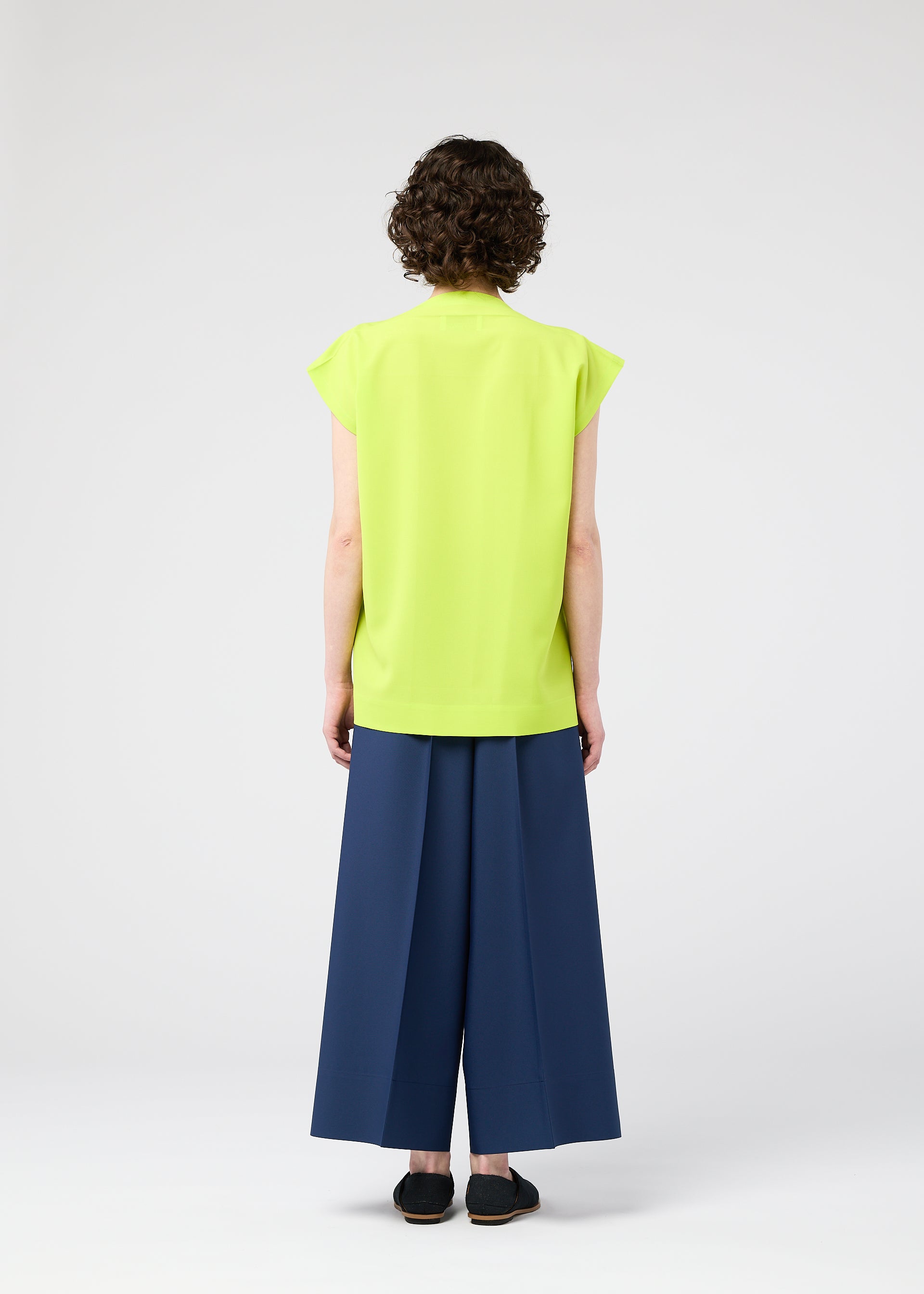 Zoomed view A person with short curly hair, seen from behind, wears the FLAT TUCK T by 132 5. ISSEY MIYAKE in bright yellow-green, styled front-tucked with wide-legged dark blue pants and black shoes against a light background for a gorgeous look.