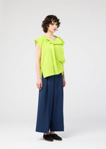 A person with short curly hair wears the FLAT TUCK T by 132 5. ISSEY MIYAKE in lime green, styled with a front tuck and paired with wide-legged navy blue pants, standing against a plain light background.