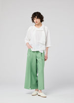 A person with short curly hair models the 132 5. ISSEY MIYAKE FLAT TUCK T, a loose white draping top with scarf detail, paired with wide-legged light green trousers and white slip-on shoes against a plain light gray background.