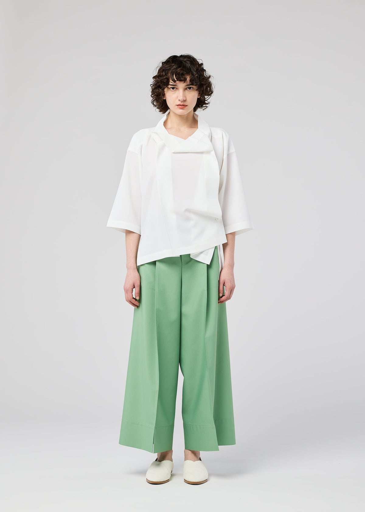 A person with curly hair stands against a light background, wearing the FLAT TUCK T by 132 5. ISSEY MIYAKE, mint green wide-legged pants, and white slip-on shoes. Their arms are by their sides and their expression is neutral. view 9 of 13