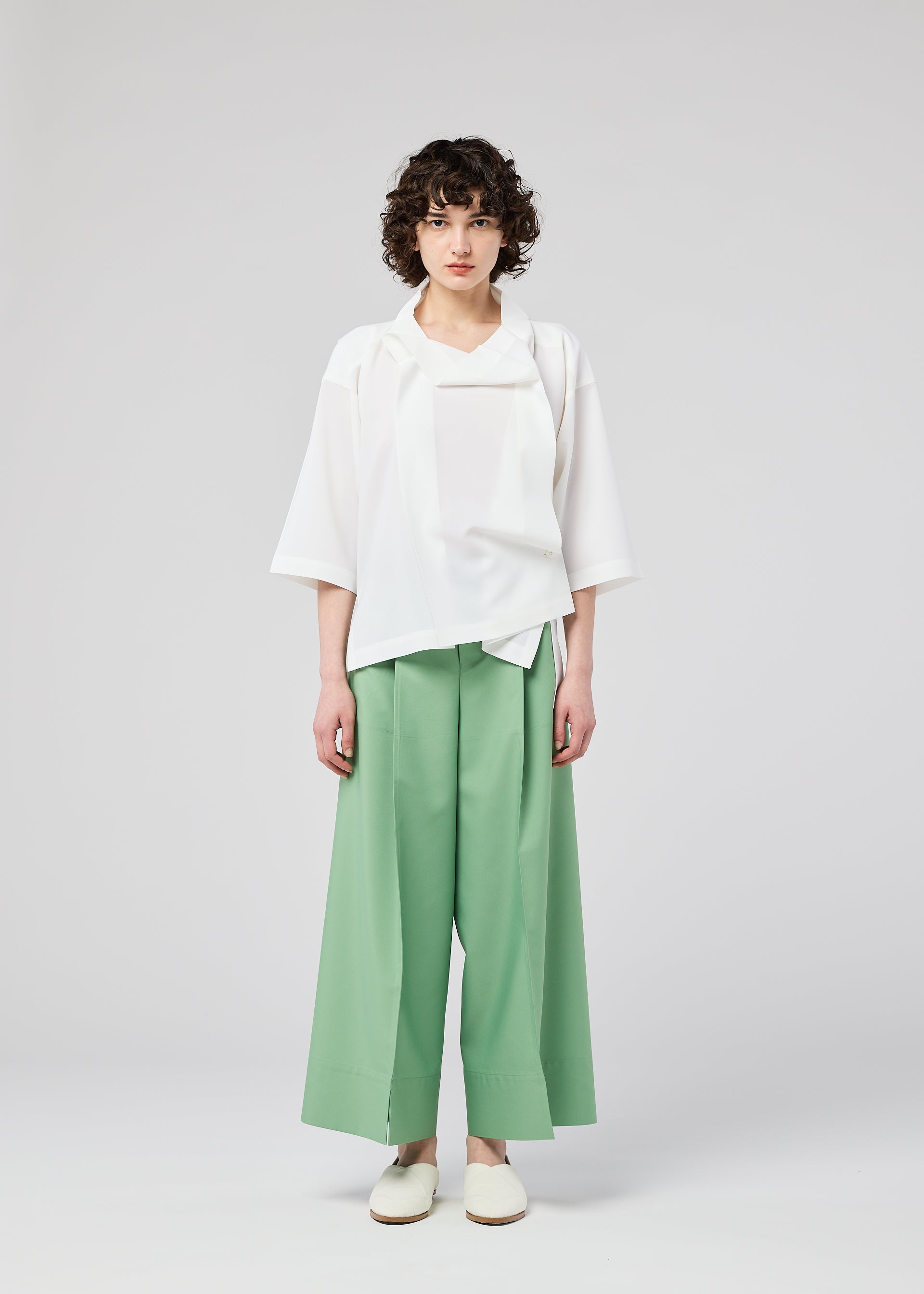 Zoomed view A person with curly hair stands against a light background, wearing the FLAT TUCK T by 132 5. ISSEY MIYAKE, mint green wide-legged pants, and white slip-on shoes. Their arms are by their sides and their expression is neutral.