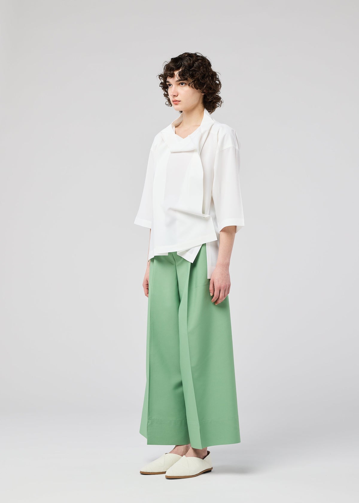 A person with short, curly hair stands against a plain light background, wearing the FLAT TUCK T by 132 5. ISSEY MIYAKE—a loose white top—paired with wide-legged mint green pants and white slip-on shoes, looking slightly to the side. view 10 of 13