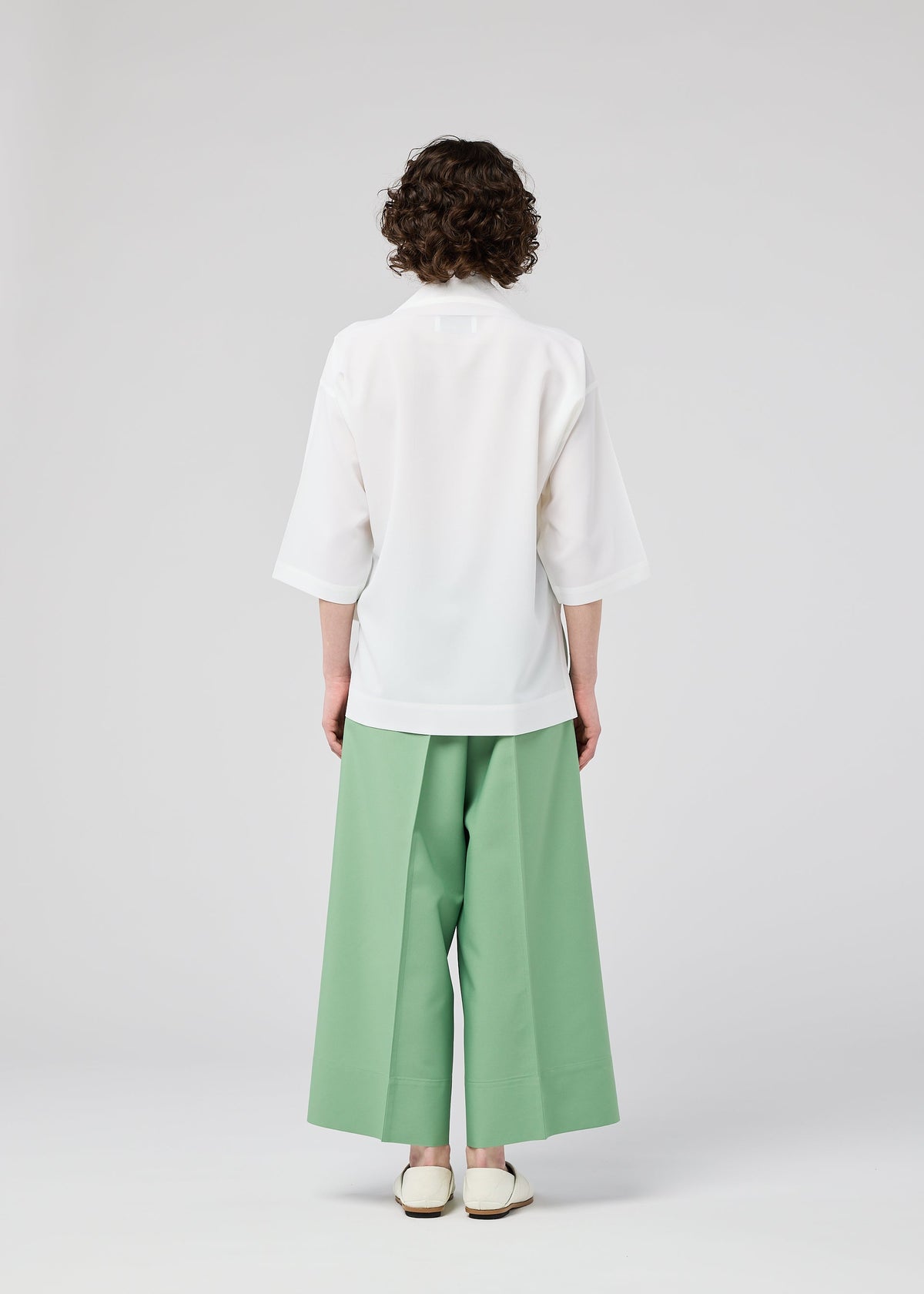 A person with short curly hair, seen from behind, wears the 132 5. ISSEY MIYAKE FLAT TUCK T—a loose white top with elbow-length sleeves—paired with wide-legged mint green pants, standing against a plain light gray background. view 11 of 13