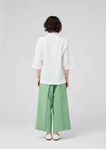 A person with short curly hair, seen from behind, wears the 132 5. ISSEY MIYAKE FLAT TUCK T—a loose white top with elbow-length sleeves—paired with wide-legged mint green pants, standing against a plain light gray background.