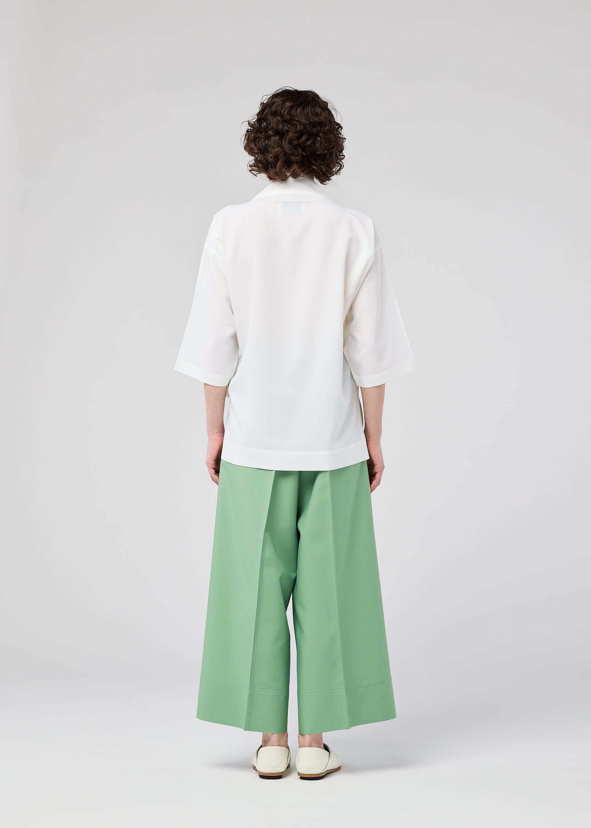 Zoomed view A person with short curly hair, seen from behind, wears the 132 5. ISSEY MIYAKE FLAT TUCK T—a loose white top with elbow-length sleeves—paired with wide-legged mint green pants, standing against a plain light gray background.
