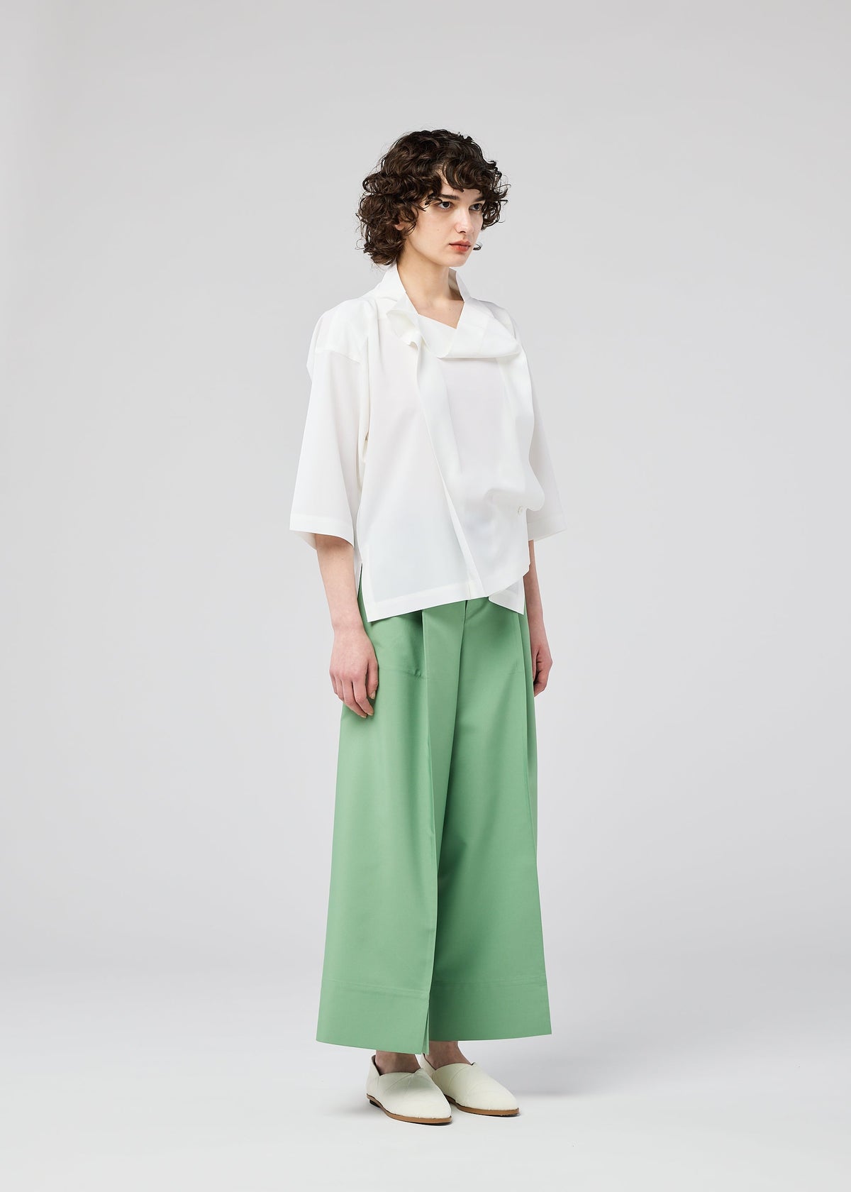 Wearing the FLAT TUCK T by 132 5. ISSEY MIYAKE, a person with short curly hair pairs it with wide green trousers and white shoes, standing against a plain light background. view 12 of 13
