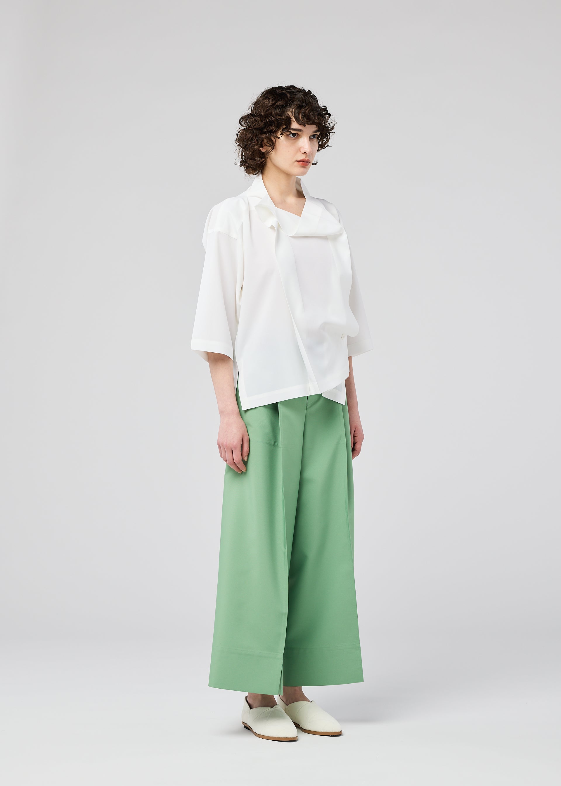 Zoomed view Wearing the FLAT TUCK T by 132 5. ISSEY MIYAKE, a person with short curly hair pairs it with wide green trousers and white shoes, standing against a plain light background.