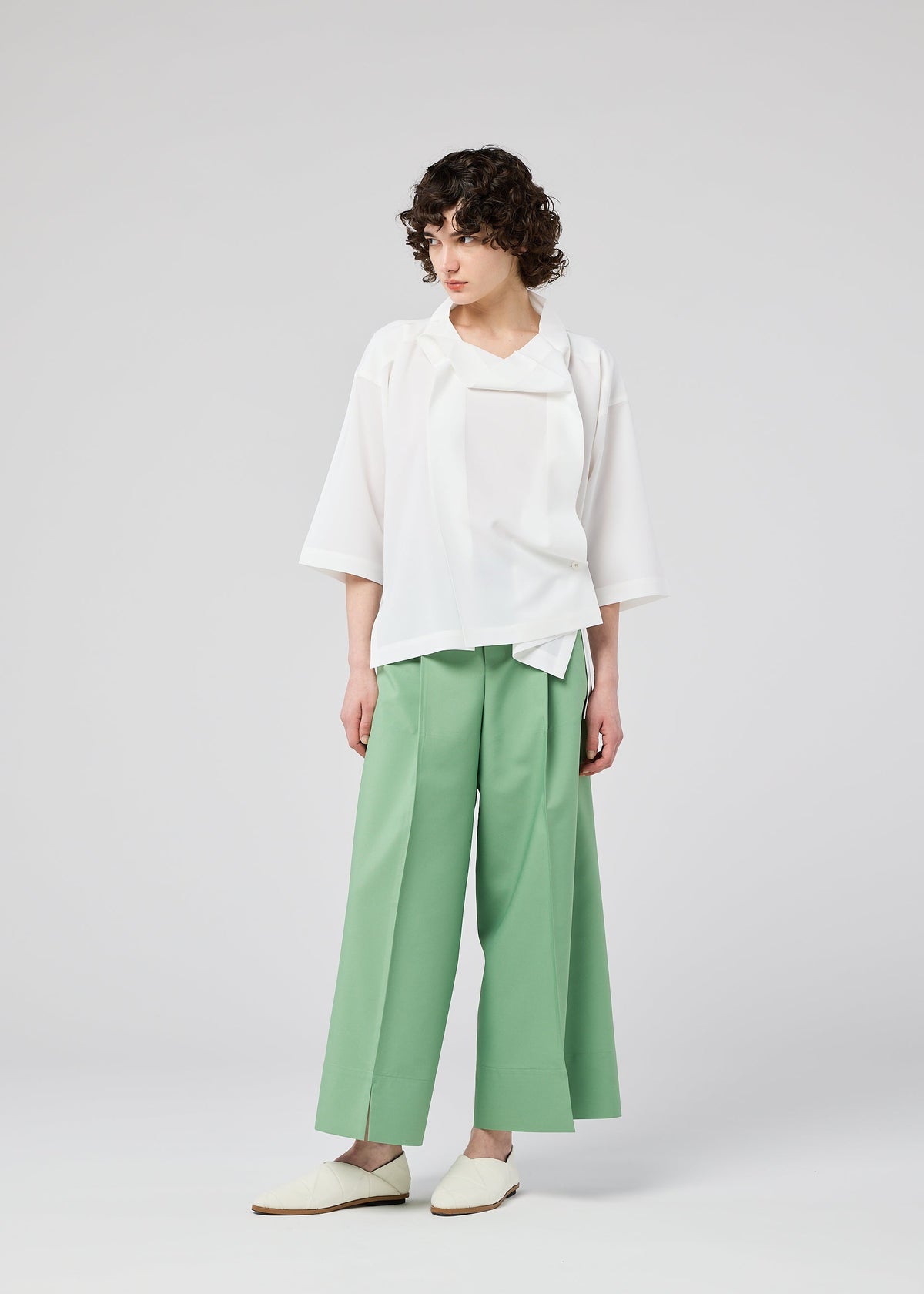 A person with short curly hair wears the FLAT TUCK T by 132 5. ISSEY MIYAKE—a loose white top with wide sleeves—and wide-legged mint green pants, paired with white shoes, standing against a plain light gray background. view 13 of 13