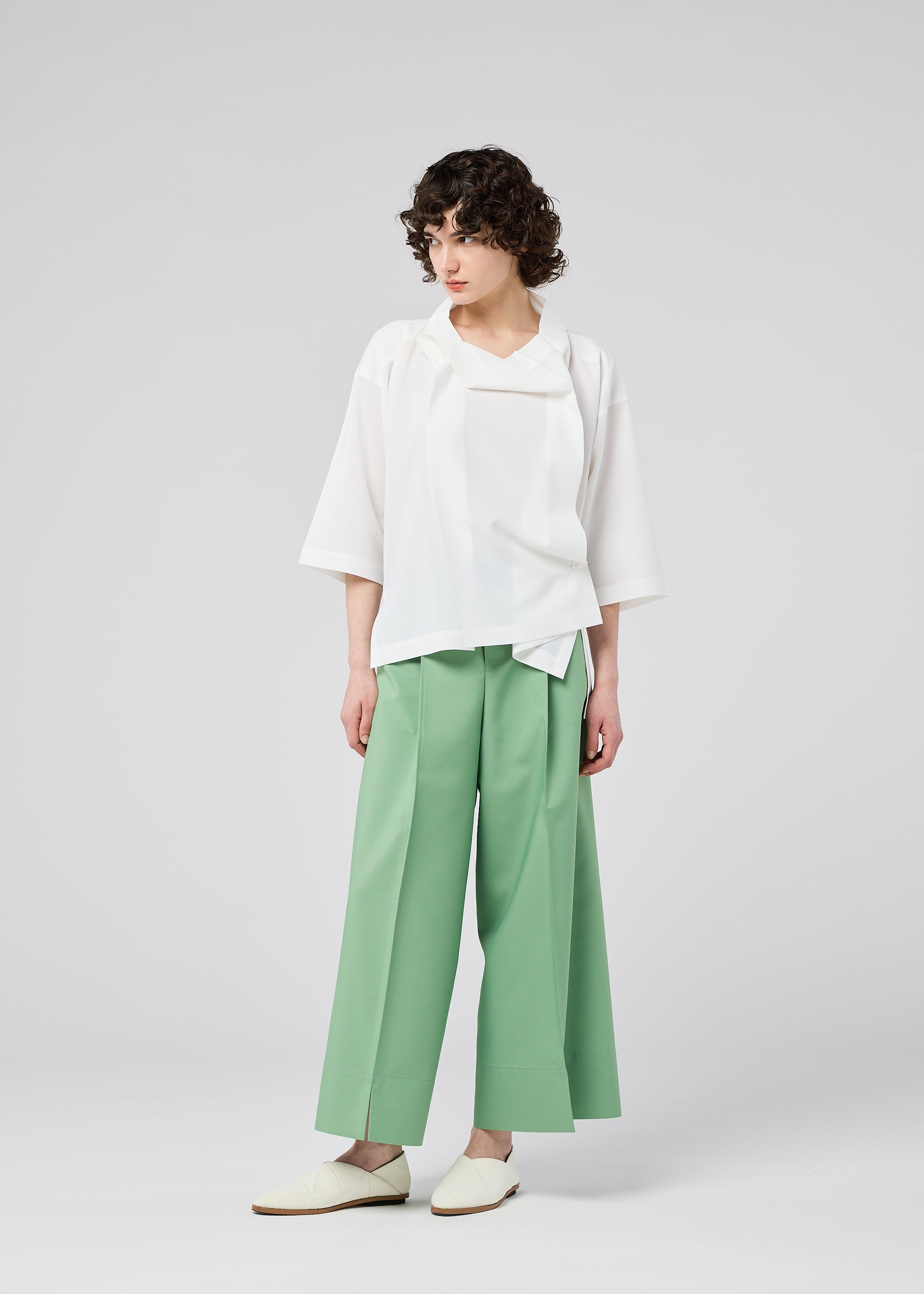 Zoomed view A person with short curly hair wears the FLAT TUCK T by 132 5. ISSEY MIYAKE—a loose white top with wide sleeves—and wide-legged mint green pants, paired with white shoes, standing against a plain light gray background.