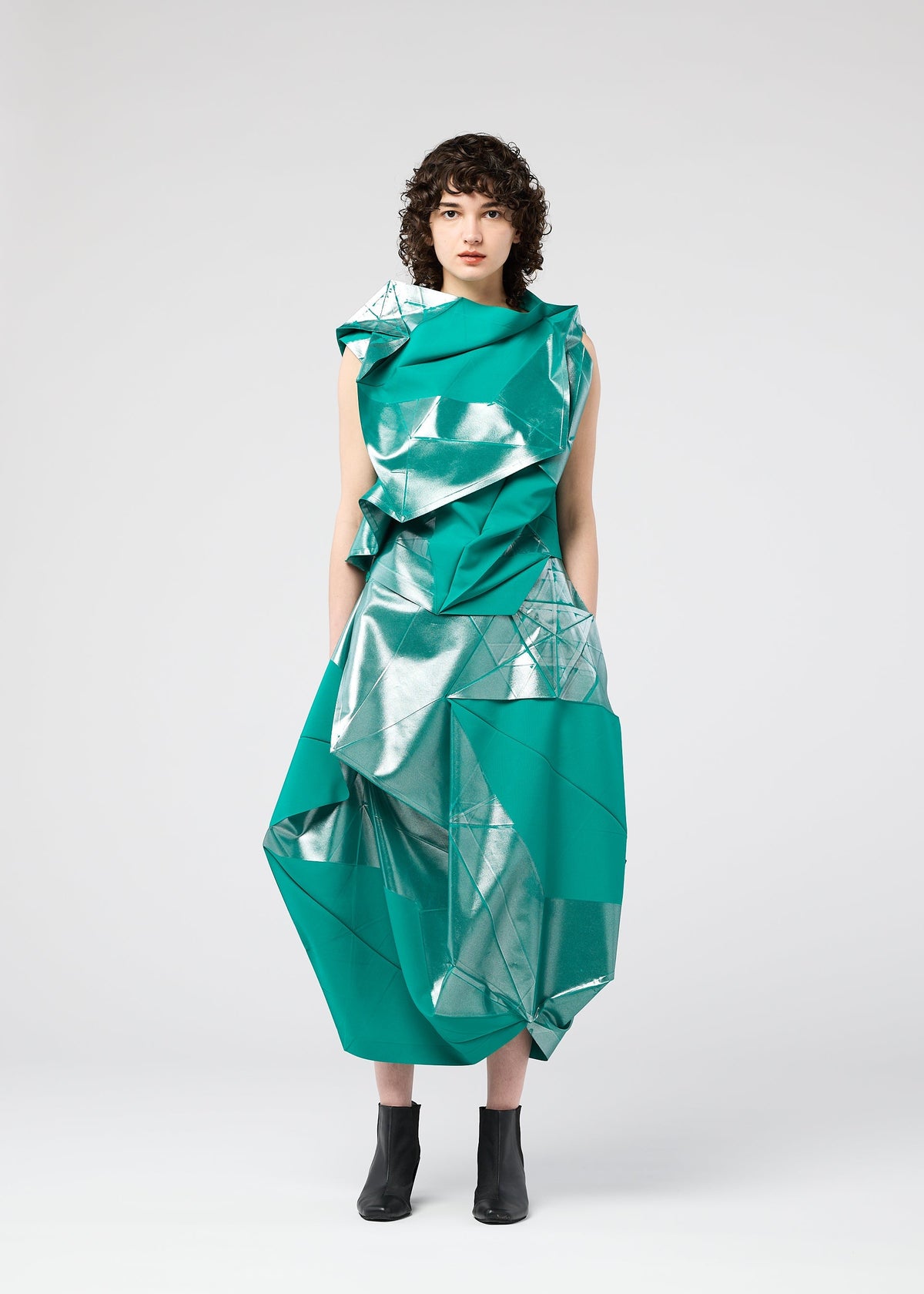 A person with short curly hair wears the 132 5. ISSEY MIYAKE NO.17 FOIL, a sculptural teal and silver metallic dress with geometric folds, against a plain light gray background, styled with black ankle boots. view 5 of 10